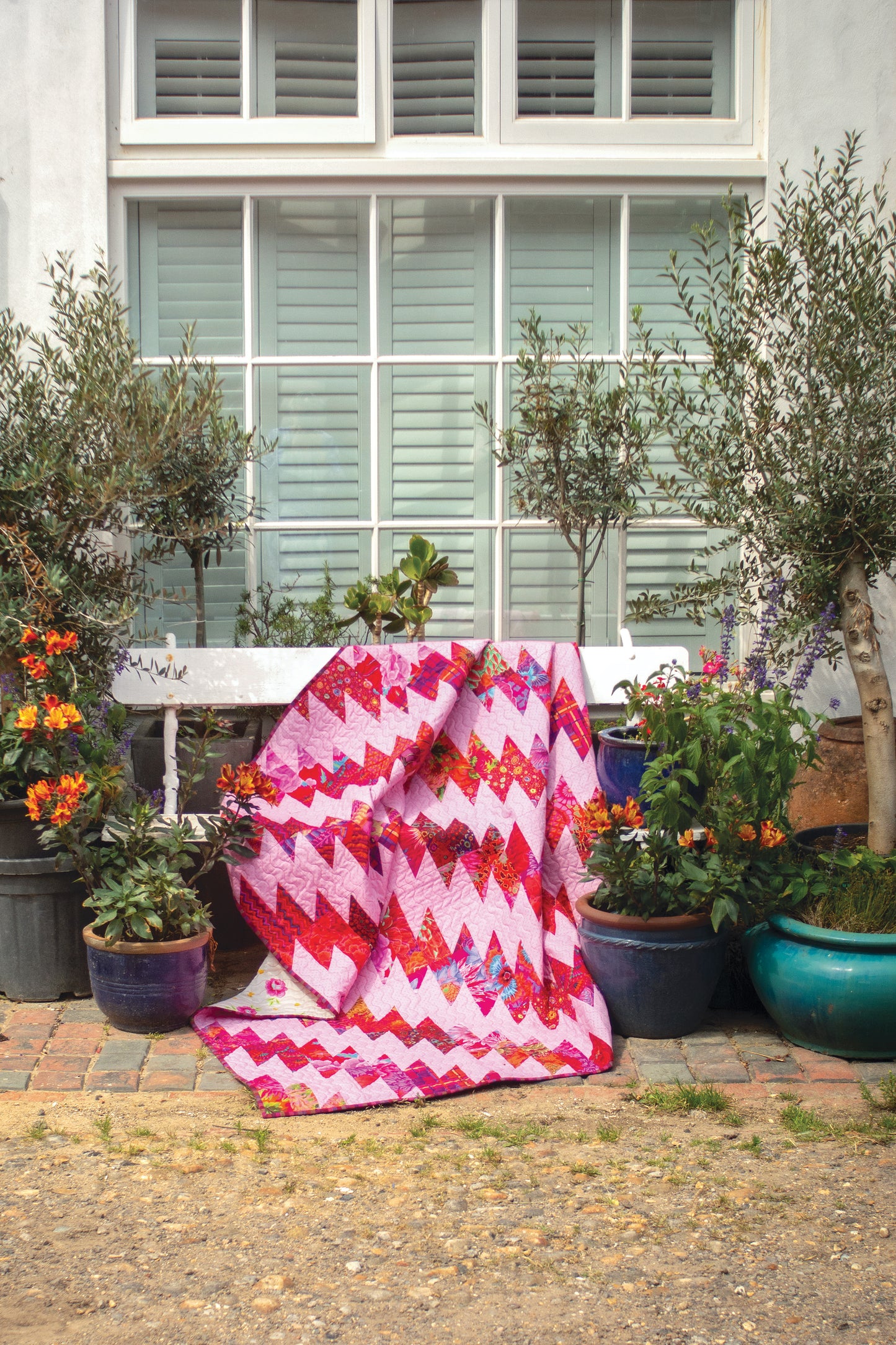 A Layer Cake Quilts creation from Stash Books, featuring a pink, red, and purple zigzag pattern with 10" precut squares, is displayed on a white garden bench amid green plants in colorful pots beside a white building with large windows.