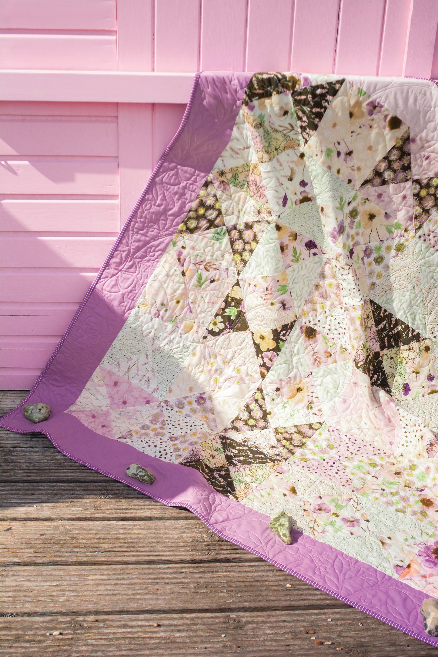 A Stash Books Layer Cake Quilts creation, made from 10" floral precut squares with a purple border, is displayed over a wooden surface before a pink wall, its corners secured by small rocks.