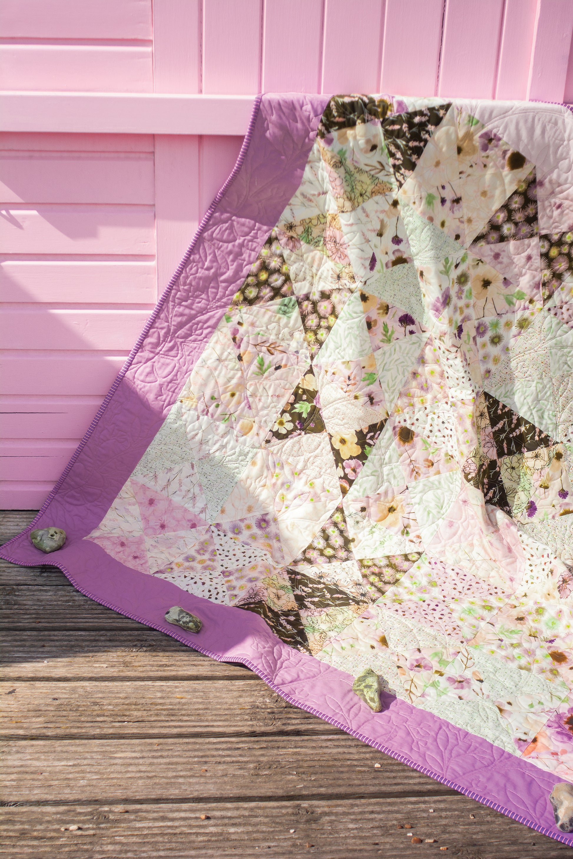 A Stash Books Layer Cake Quilts creation, made from 10" floral precut squares with a purple border, is displayed over a wooden surface before a pink wall, its corners secured by small rocks.