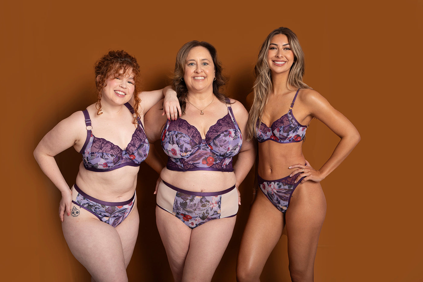 Three women of varied body types and skin tones smile together in matching purple floral lingerie, highlighting the craftsmanship promoted in "Sew Underwire Bras & Corsets" by Stash Books, all set against a plain brown backdrop.