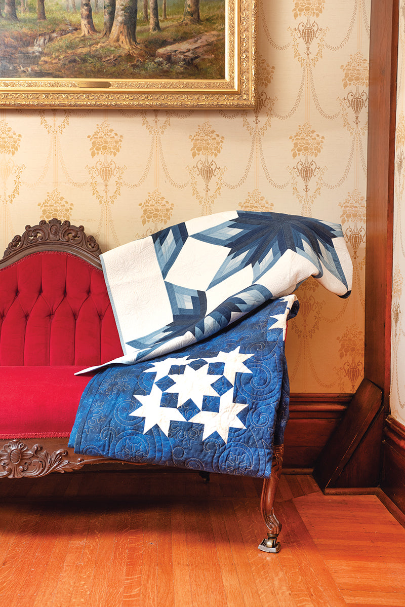 A red velvet antique sofa on a wooden floor is draped with two blue and white “Elm Creek Quilts Christmas” quilts by C&T Publishing. A framed landscape painting hangs above the sofa on patterned wallpaper.