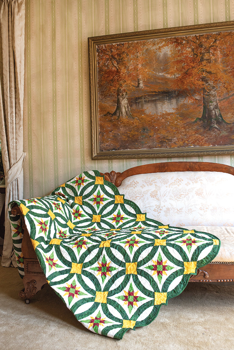 A vintage wooden sofa with pale floral upholstery is draped with the C&T Publishing Elm Creek Quilts Christmas quilt, showcasing green, yellow, and red star and ring designs. Above it, a large autumn landscape painting hangs on the wallpapered wall.