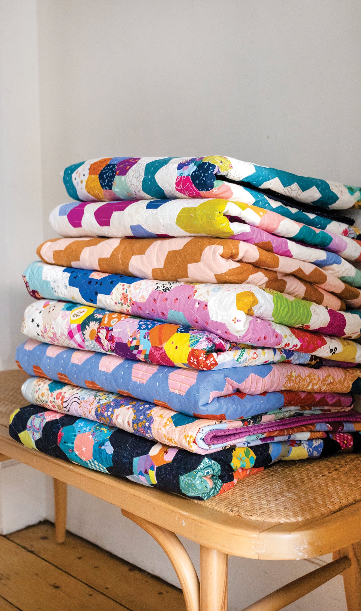A tall stack of colorful, English paper piecing 50 Hexagon Quilts from Stash Books sitting on a wooden chair.