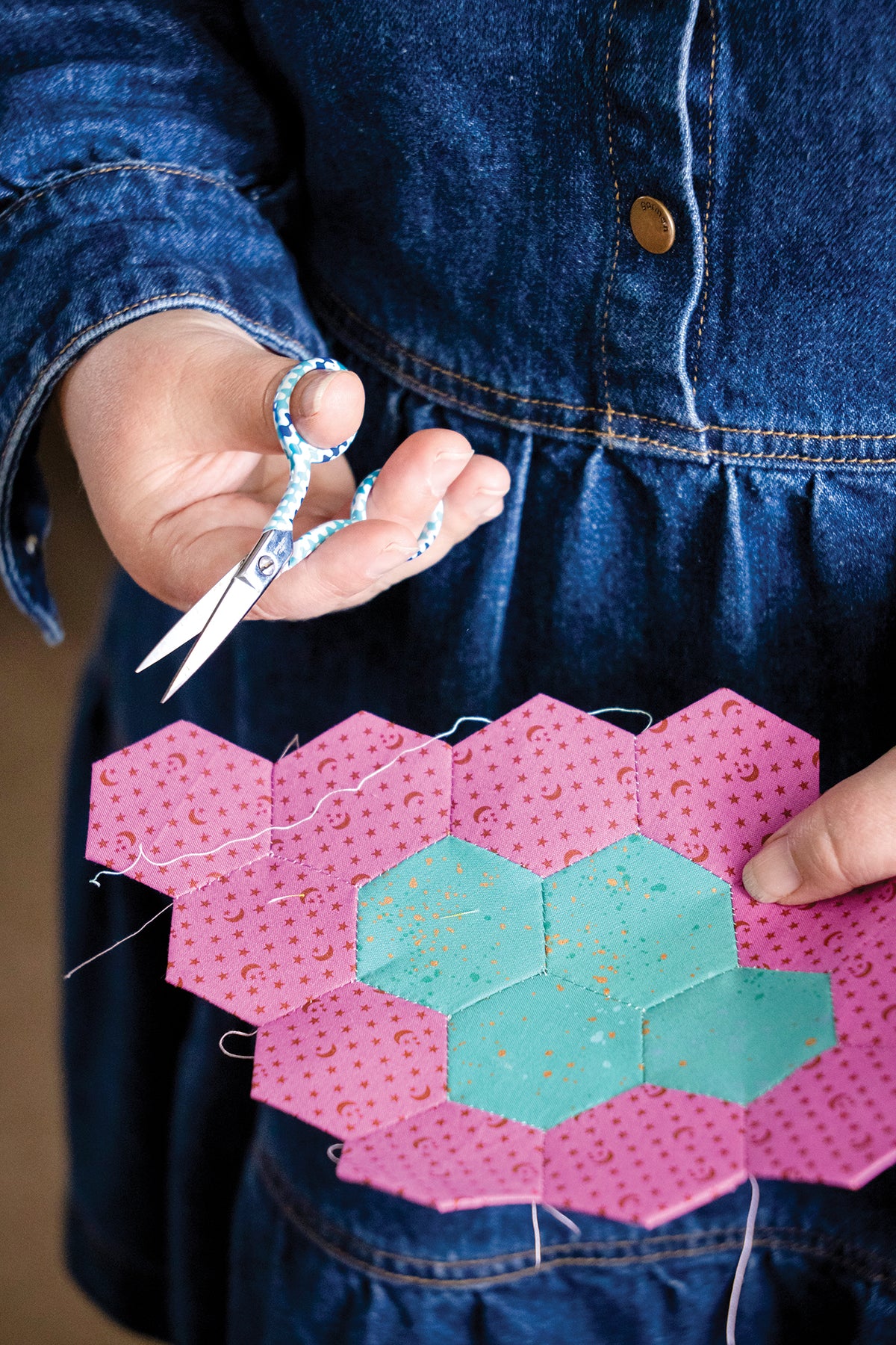 Hands holding small scissors and a piece of fabric featuring an English paper piecing design of pink and teal hexagons from 50 Hexagon Quilts.