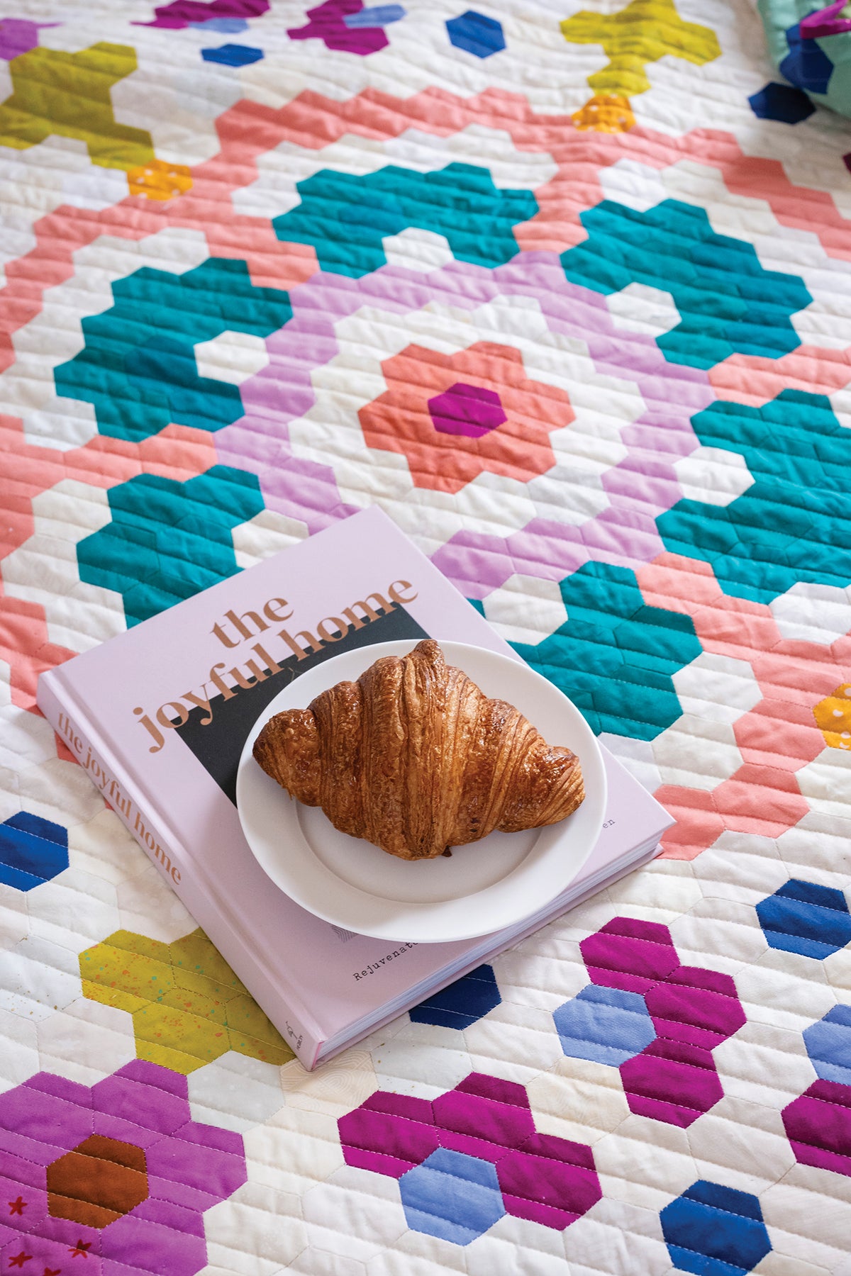 A croissant on a small plate rests on a book titled The Joyful Home, which is placed on a colorful, geometric 50 Hexagon Quilts quilt.