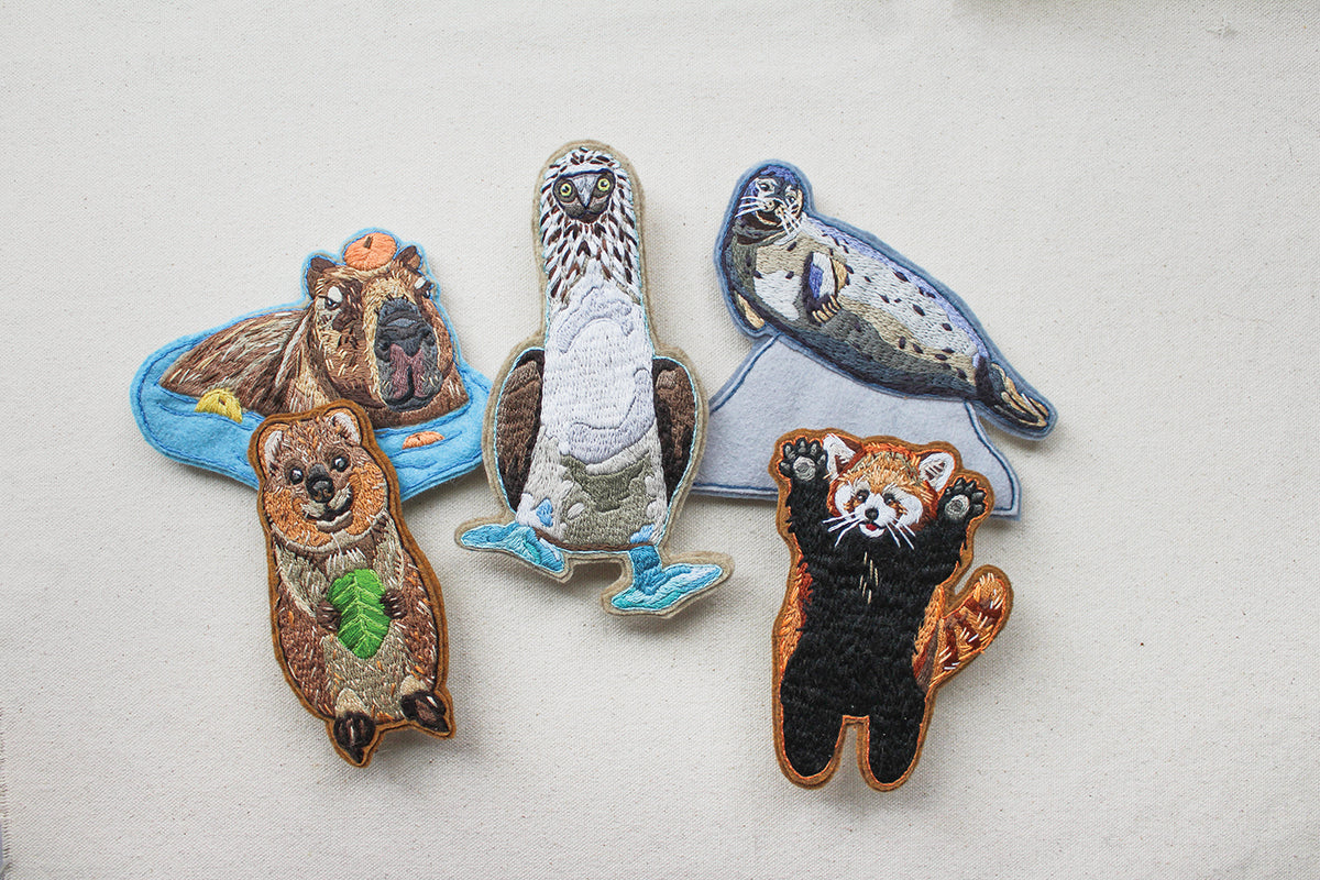 Five Wild & Wearable Animal Embroidery accessories featuring embroidered patches of a capybara, quokka, blue-footed booby, spotted seal, and red panda.