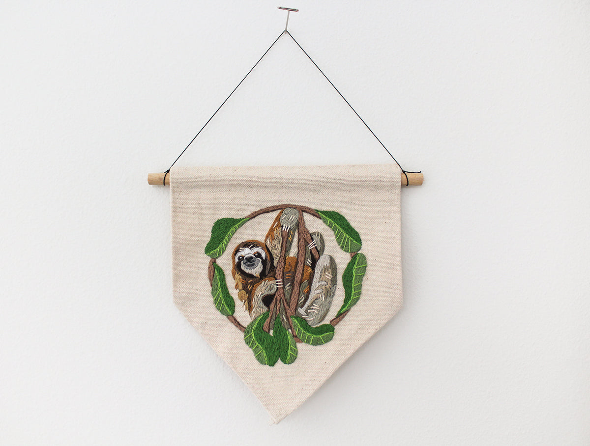 Wild & Wearable Animal Embroidery fabric wall hanging featuring a sloth dangling from a branch encircled by leaves.
