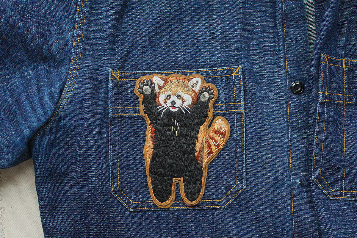 An embroidered patch of a red panda with its arms raised, stitched onto the pocket of a denim shirt as a piece of Wild & Wearable Animal Embroidery.