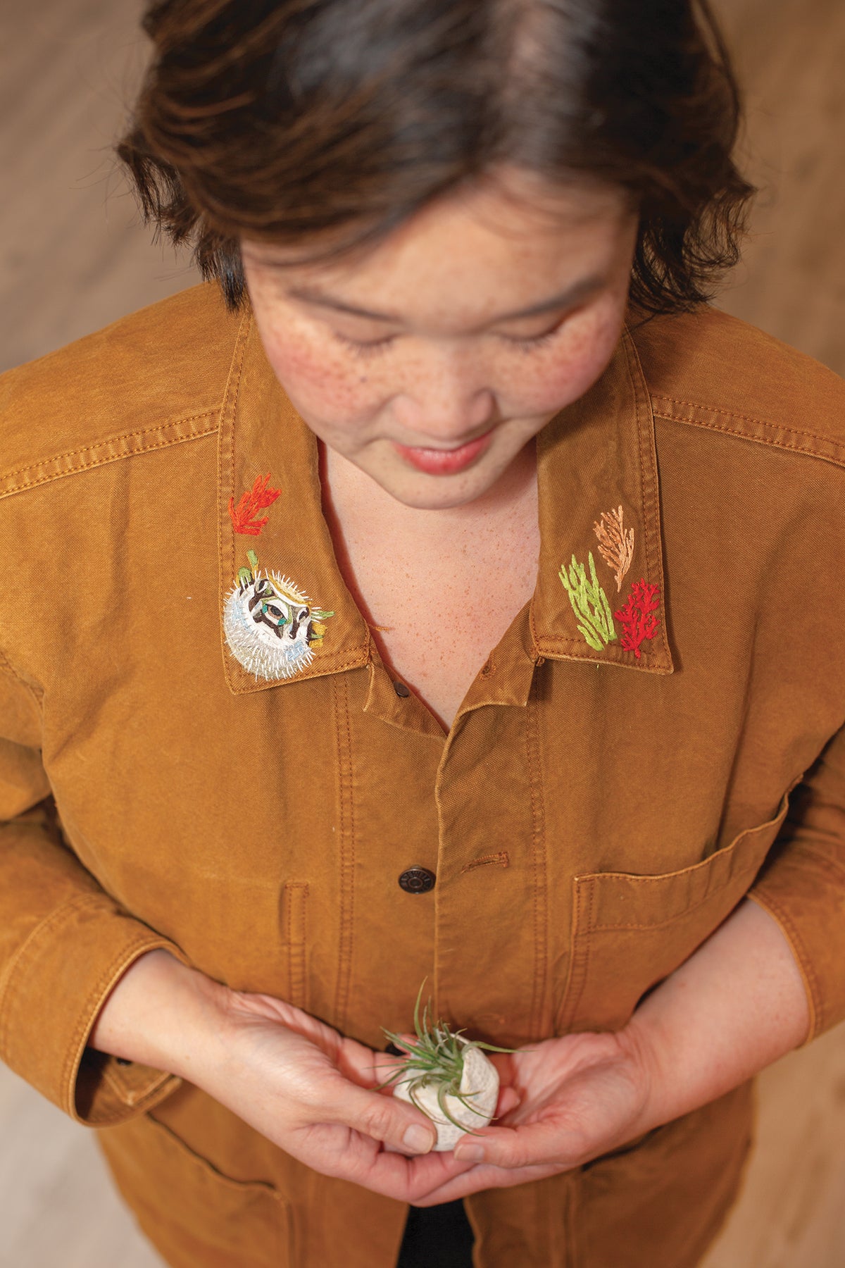 A person wearing a brown Wild & Wearable Animal Embroidery jacket with handmade coral and pufferfish collar details, holding a small air plant in their cupped hands.