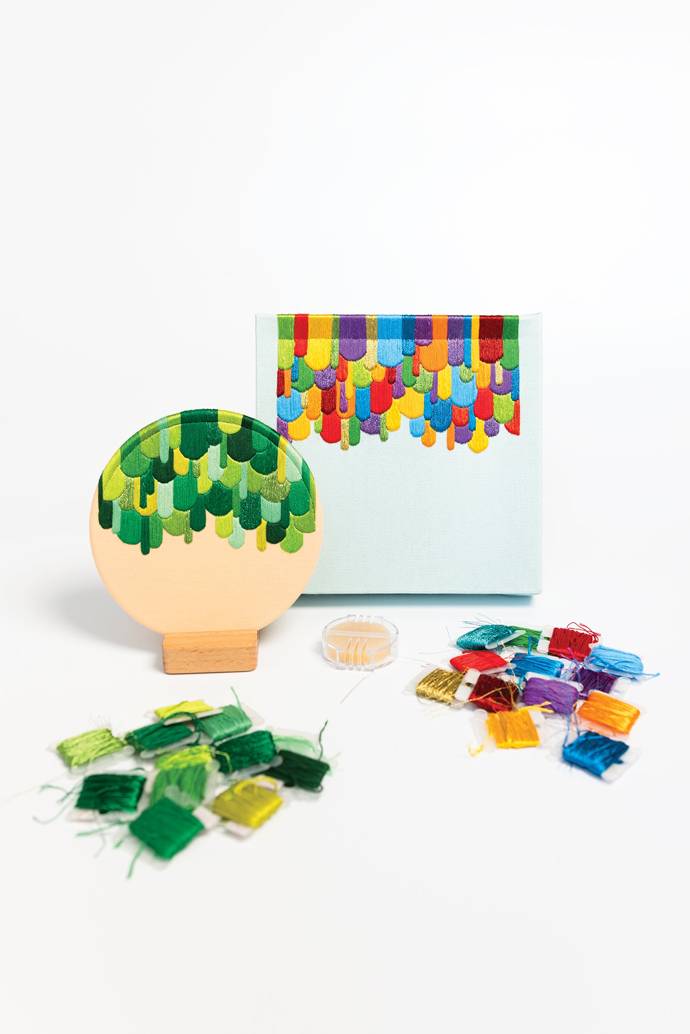 The Modern Embroidery, A Field Guide by Stash Books features a round embroidery hoop with green, yellow, and blue stitches displayed on a sturdy wooden base. Ideal for beginner embroidery projects, it comes with colorful threads and a blue canvas coordinated with matching stitches, all beautifully arranged on a clean white background.