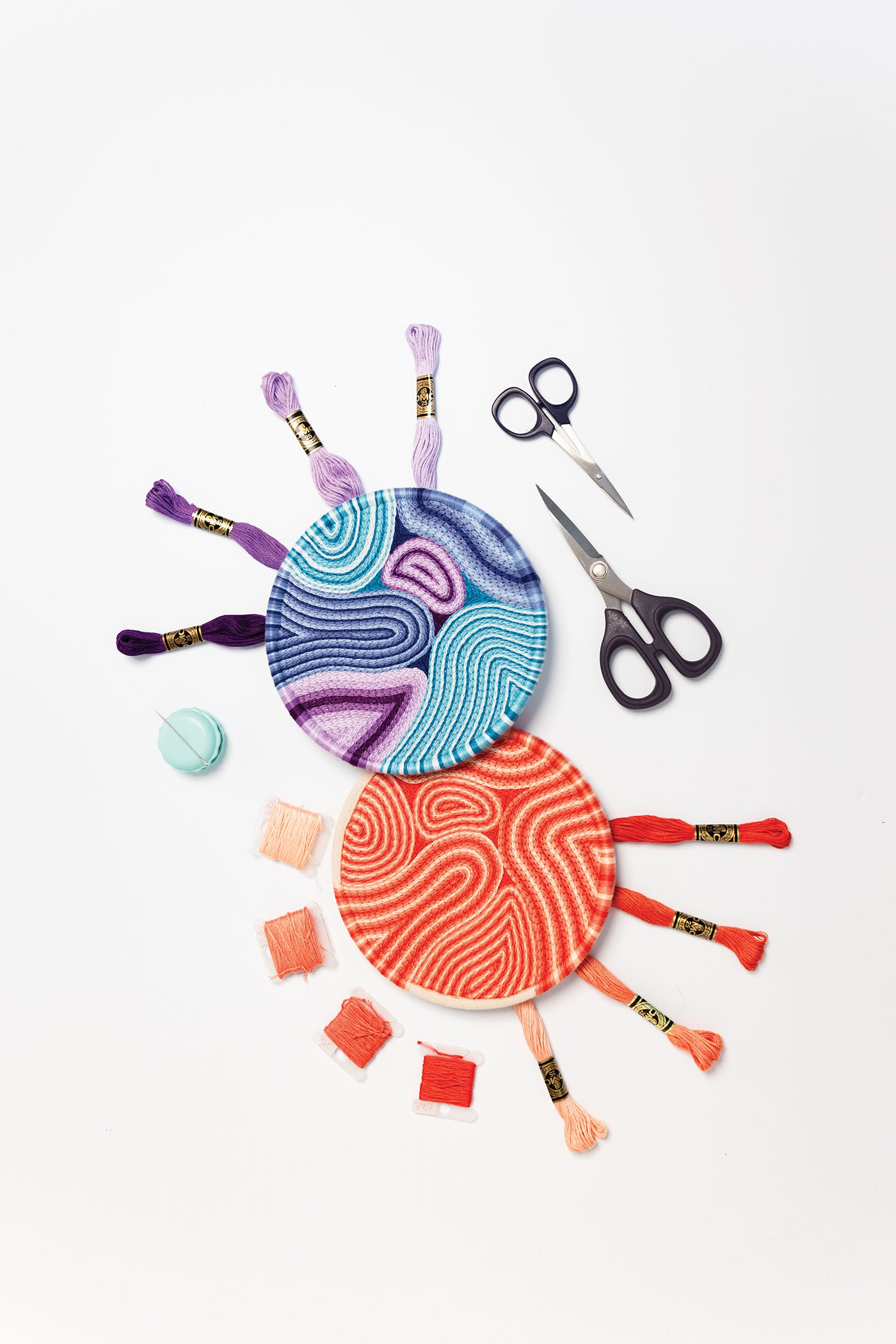 Two round plates with colorful threaded patterns, surrounded by embroidery threads, scissors, a needle threader, and a few spools, all arranged neatly on a white background—an ideal setup for anyone inspired by _Modern Embroidery, A Field Guide_ from Stash Books and searching for easy-to-follow beginner instruction.