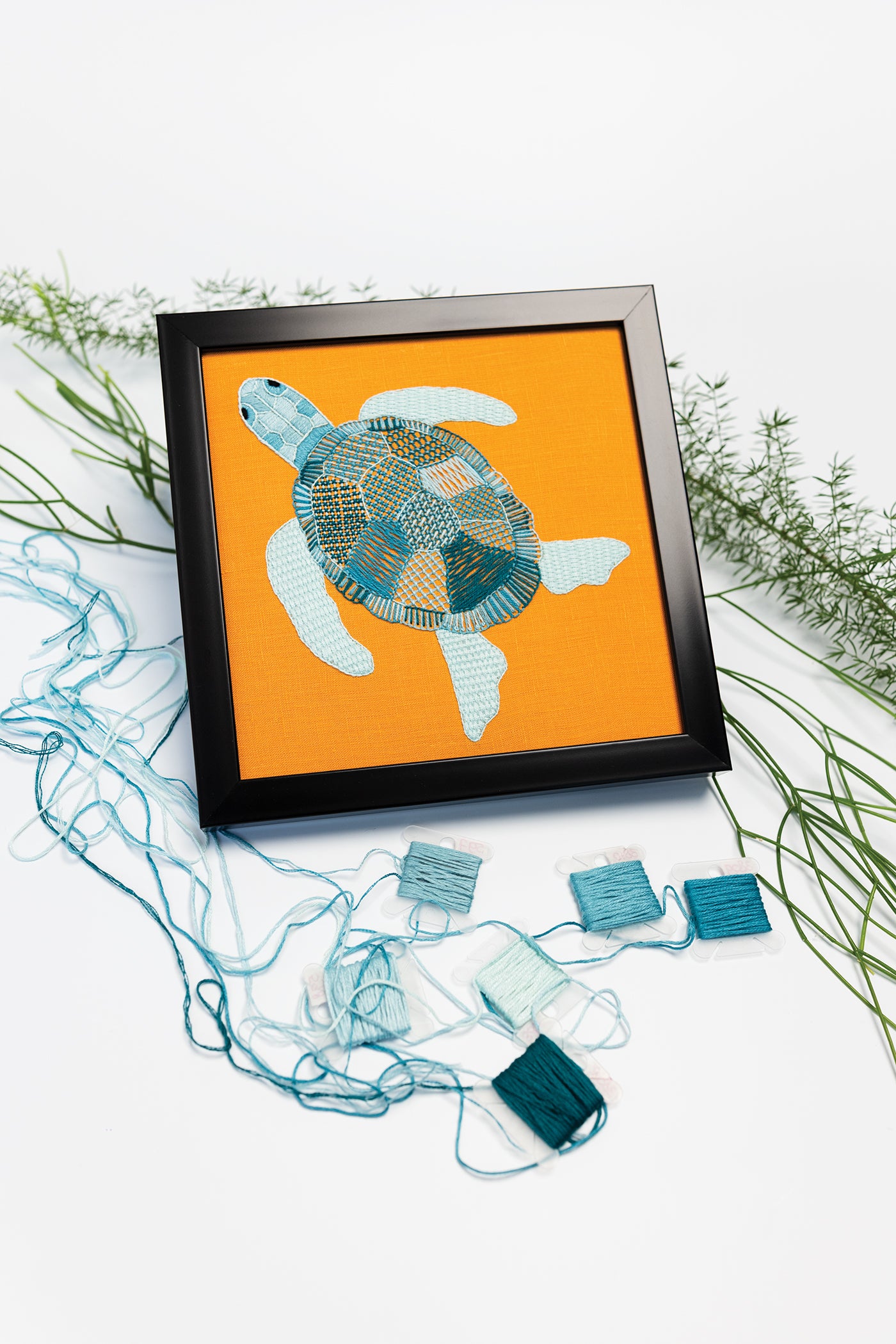 A framed turtle embroidery on orange fabric, featured in “Modern Embroidery, A Field Guide” by Stash Books, showcases a variety of embroidery stitches and is surrounded by green foliage and blue embroidery threads on a white background.