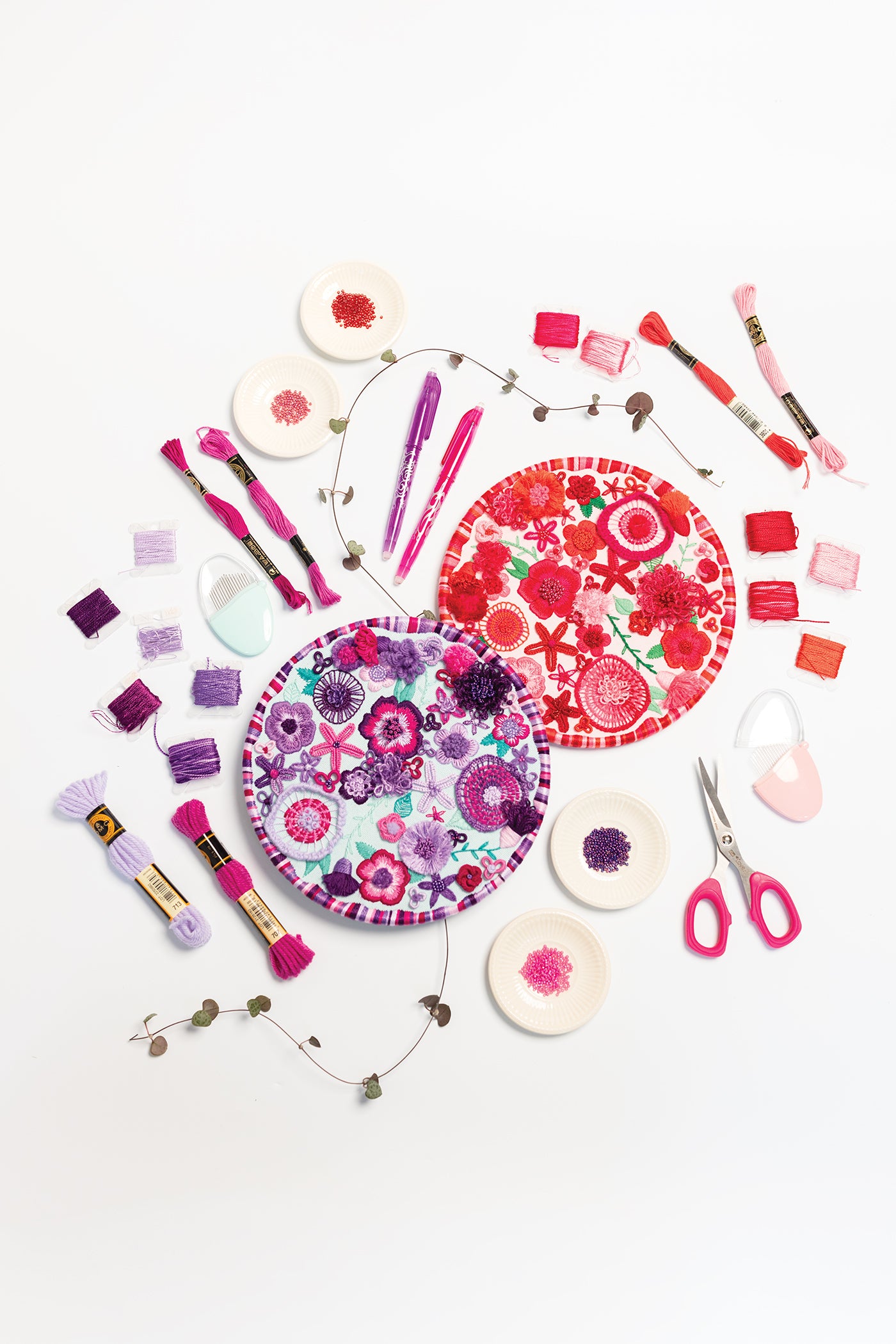 Two vibrant circular paper plates featuring floral designs are surrounded by embroidery threads, pens, small scissors, bead-filled plates, and colorful fabric swatches—all arranged in pink, purple, and red tones atop a white background. This lively scene is the perfect inspiration for anyone interested in exploring embroidery with Modern Embroidery, A Field Guide by Stash Books—an ideal resource for learning beginner embroidery stitches or finding creative ideas.