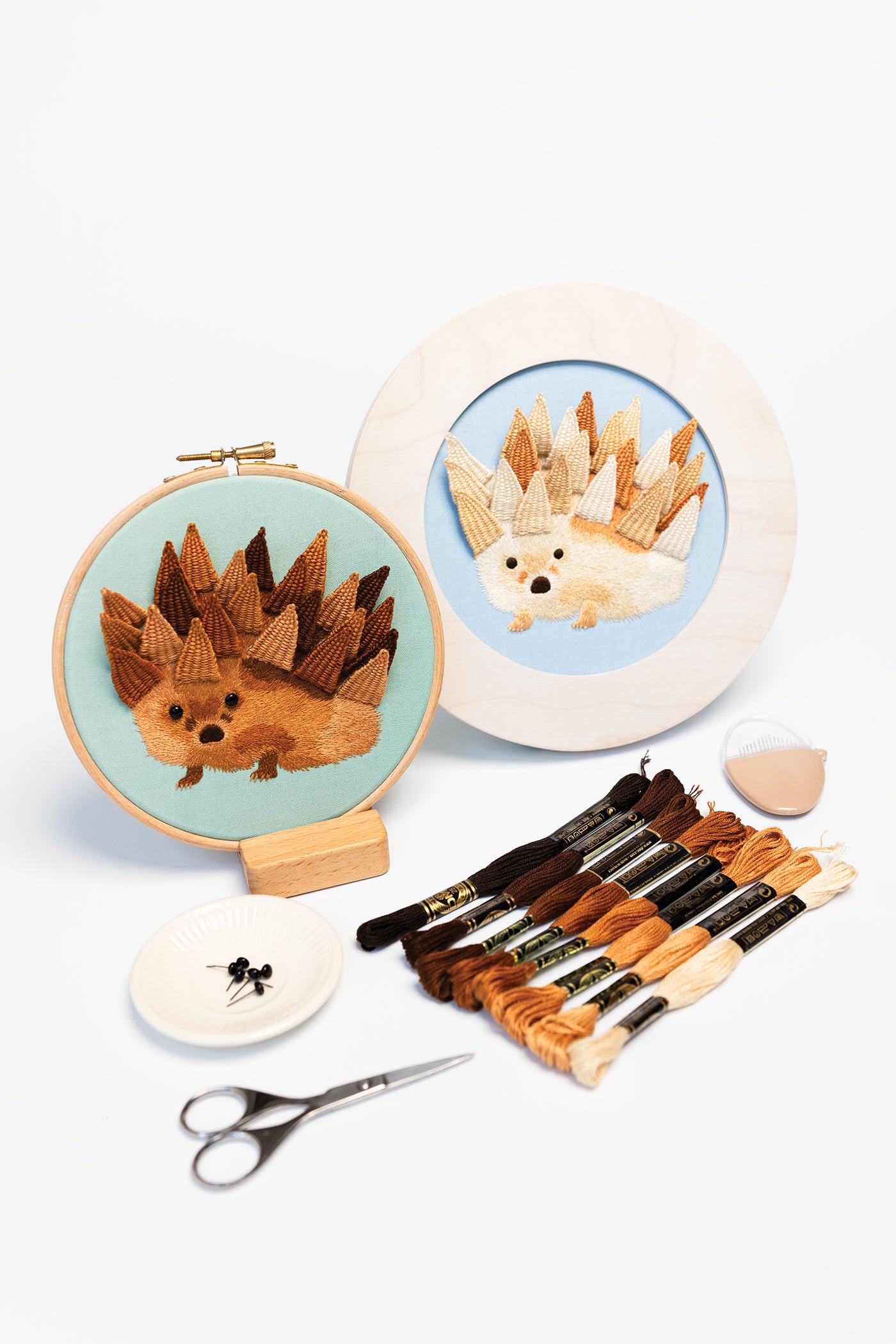 Two embroidery hoops featuring hedgehog designs, brown thread skeins, scissors, black beads in a dish, and a seashell-shaped needle holder are arranged on a white surface—an inviting setup for anyone inspired to explore beginner embroidery with Modern Embroidery, A Field Guide by Stash Books.