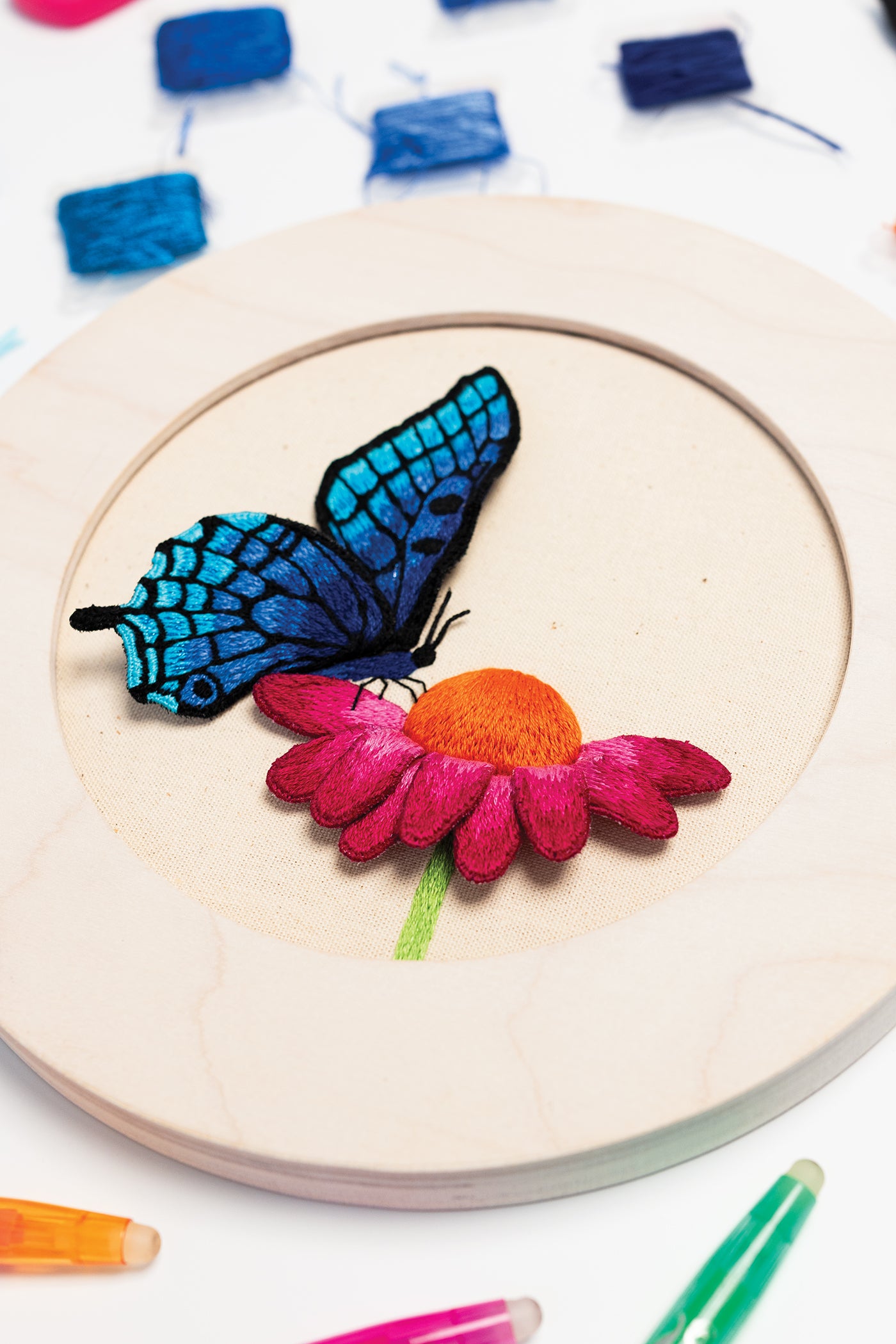 The Modern Embroidery, A Field Guide by Stash Books features colorful embroidery hoop art depicting a blue butterfly resting on a pink flower with an orange center, all set against beige fabric. Surrounded by vibrant threads and pencils, this piece offers charming inspiration for beginners or anyone looking to explore new embroidery stitches.