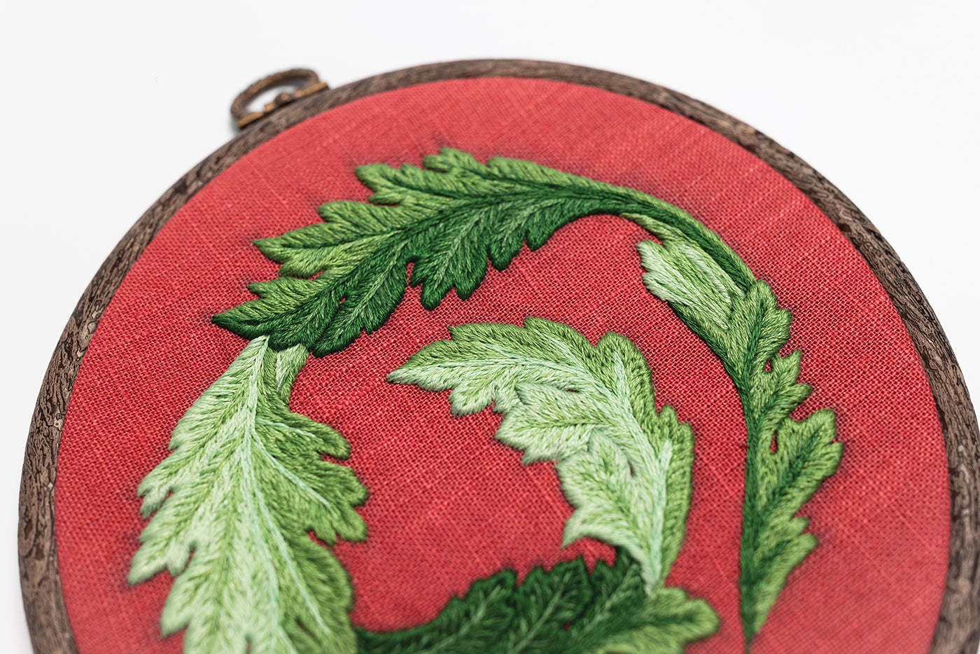 A close-up of green embroidered leaves arranged in a circular pattern on red fabric, displayed in a round wooden embroidery hoop—a perfect inspiration for beginner embroidery enthusiasts exploring new stitches, as featured in “Modern Embroidery, A Field Guide” by Stash Books.