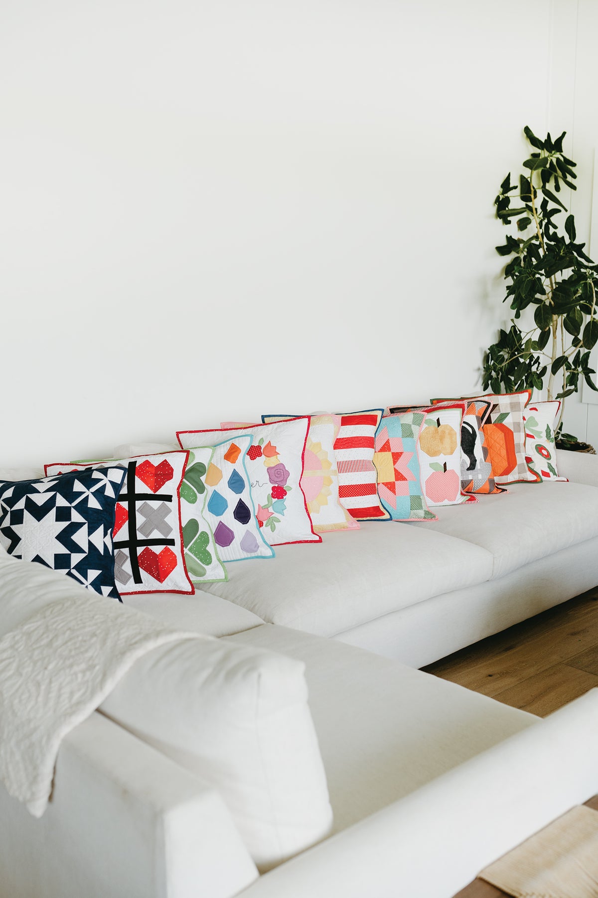 A row of twelve decorative throw pillows from Sew a Month with assorted colorful patchwork designs from Riley Blake Designs arranged on a white sofa.
