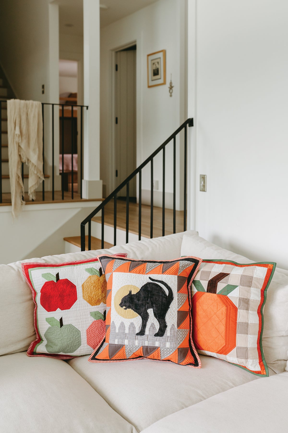 Three Sew a Pillow a Month quilted pillows featuring apple, black cat, and pumpkin designs arranged on a white sofa.
