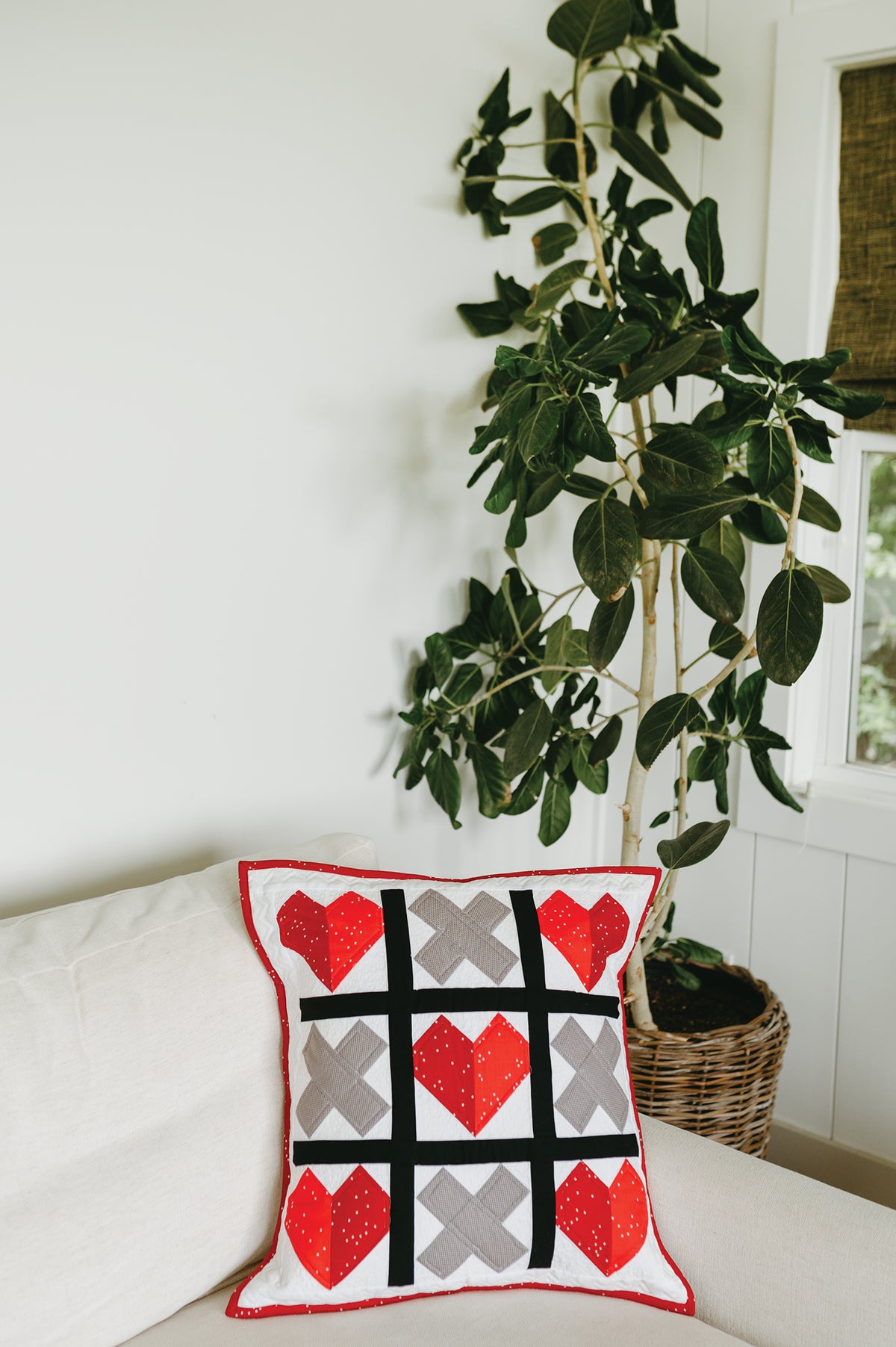 A Sew a Pillow a Month decorative throw pillow on a white sofa, featuring a quilted tic-tac-toe board pattern with red heart and grey X shapes, perfect for seasonal decorating.