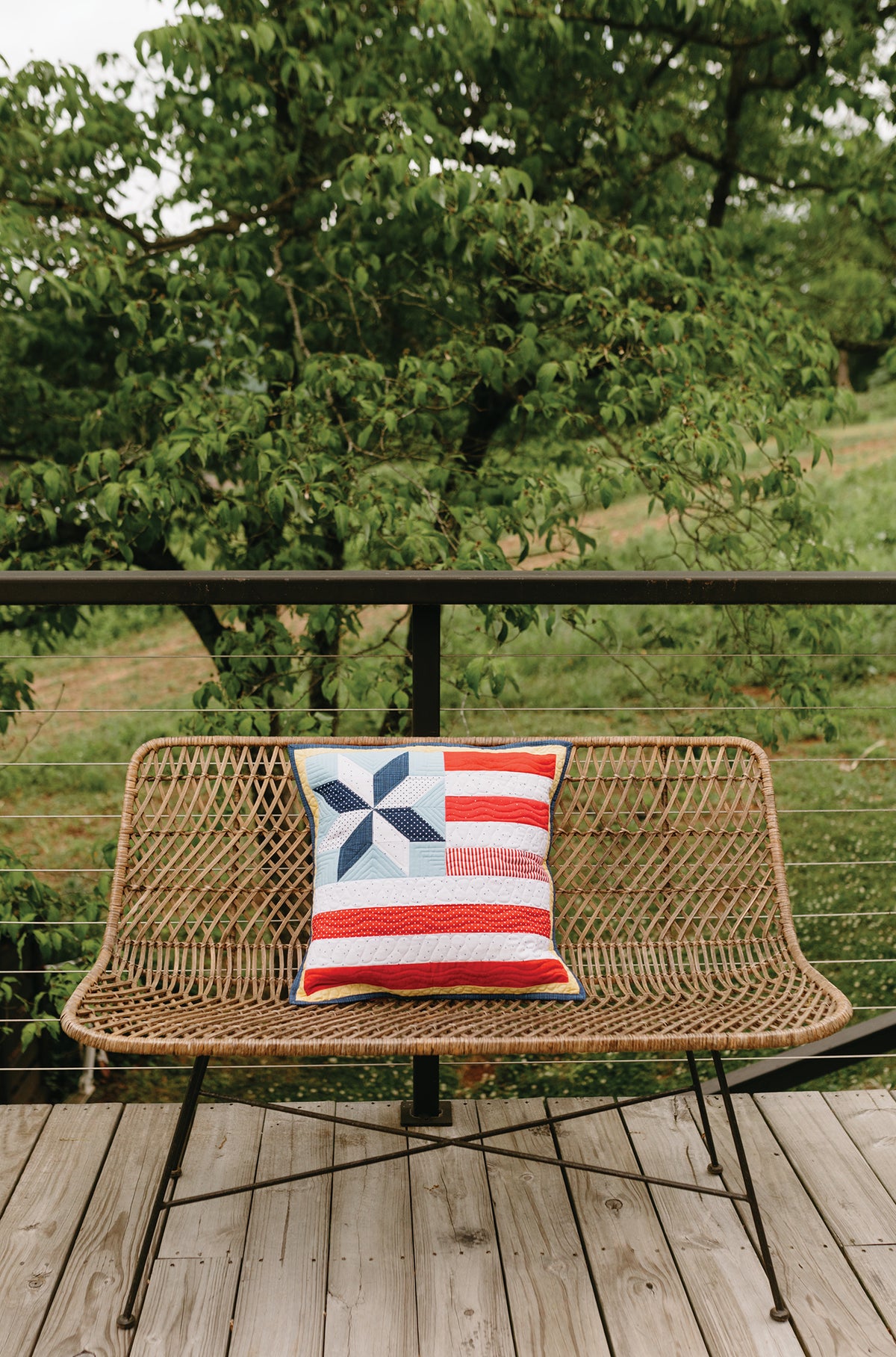 A square quilted pillow from the Sew a Month project with a blue star and red, white, and blue stripes sitting on an outdoor woven bench for seasonal decorating.