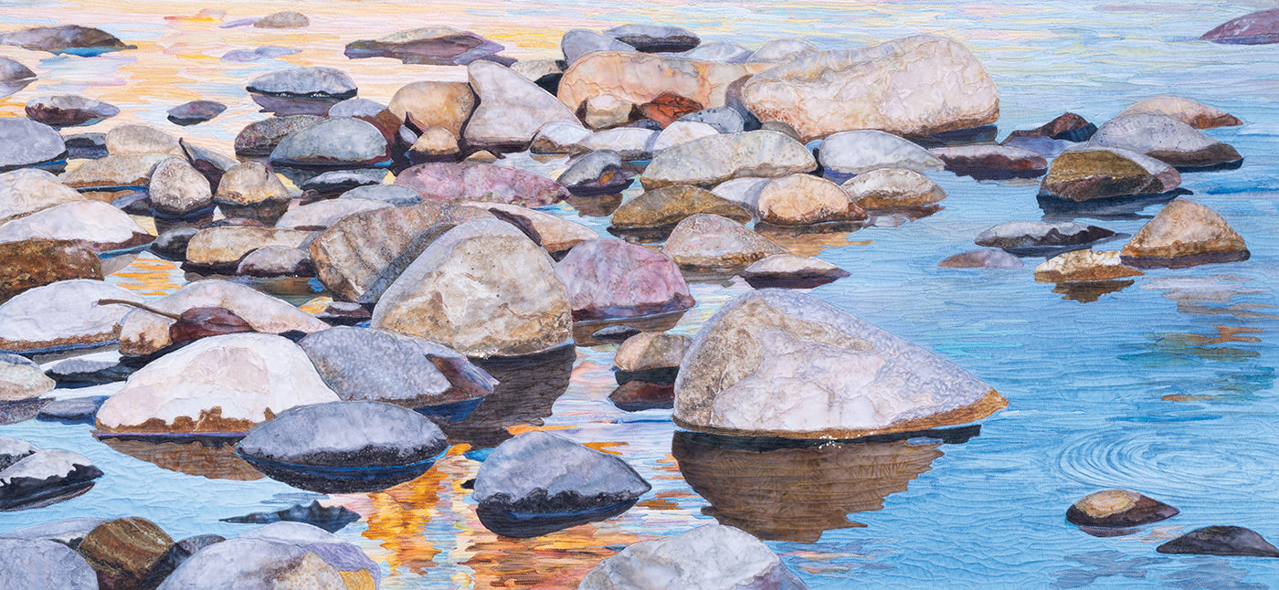 Shallow, serene waters near the shore reflect a vibrant blue sky and golden sunlight upon large, rounded rocks, artistically rendered with the textured detail of an art quilt in *Quilted Realism* by C&T Publishing.