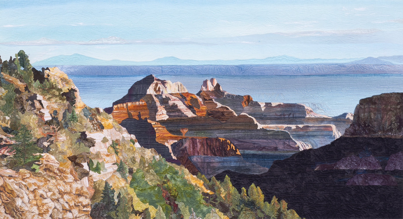 The “Quilted Realism” art quilt from C&T Publishing features a colorful, textured landscape with rocky cliffs and green trees above a vast blue lake, distant mountains, and a pale sky—beautifully capturing the essence of nature with striking quilted detail.