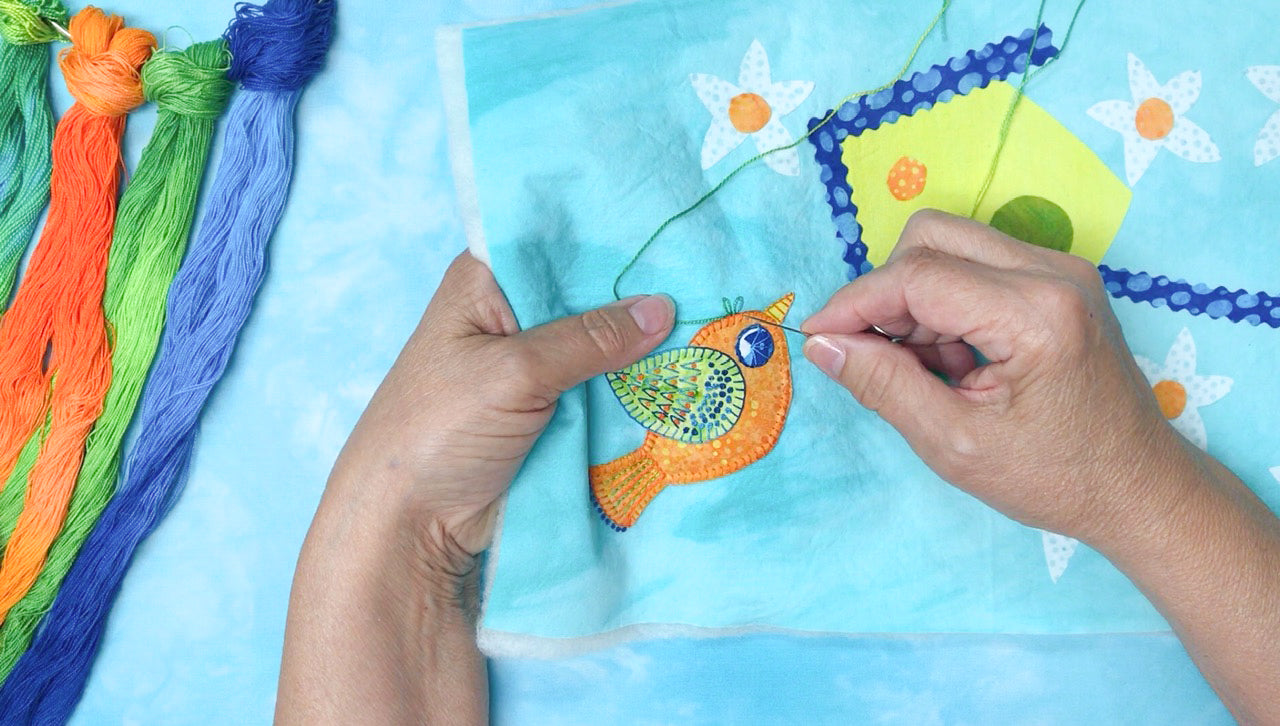 A person hand-stitching the “Fuse with Flair: Little Bird and Daisies with Stitch Library test” by Creative Spark onto light blue fabric, with an array of green, orange, blue, and purple embroidery threads visible on the left side.