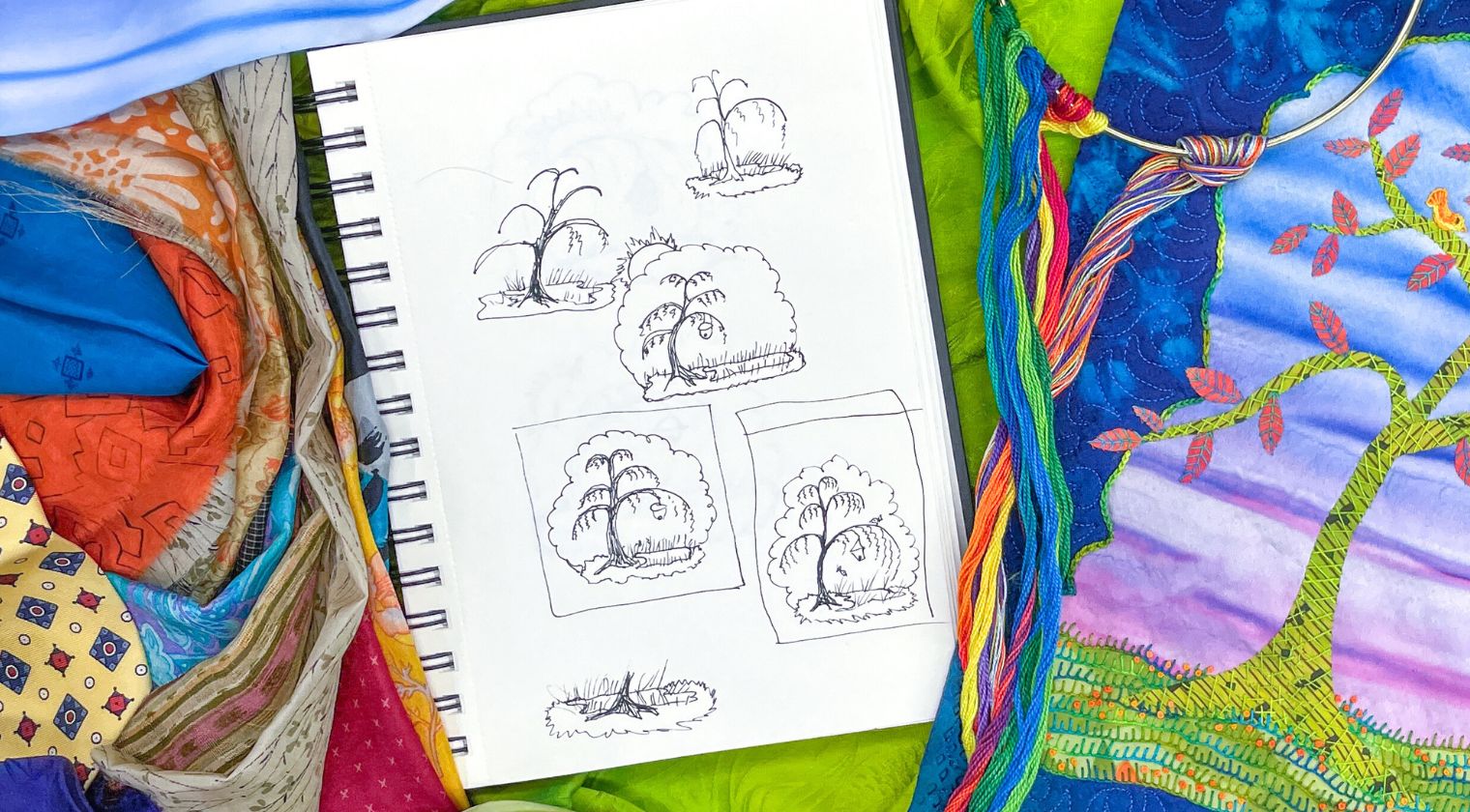 A sketchbook with tree and bush doodles lies on colorful fabric alongside Creative Spark’s “Silk Art Quilts Made Simple with Stitch Library,” a quilted artwork featuring intricate hand embroidery, with embroidery floss draped nearby.