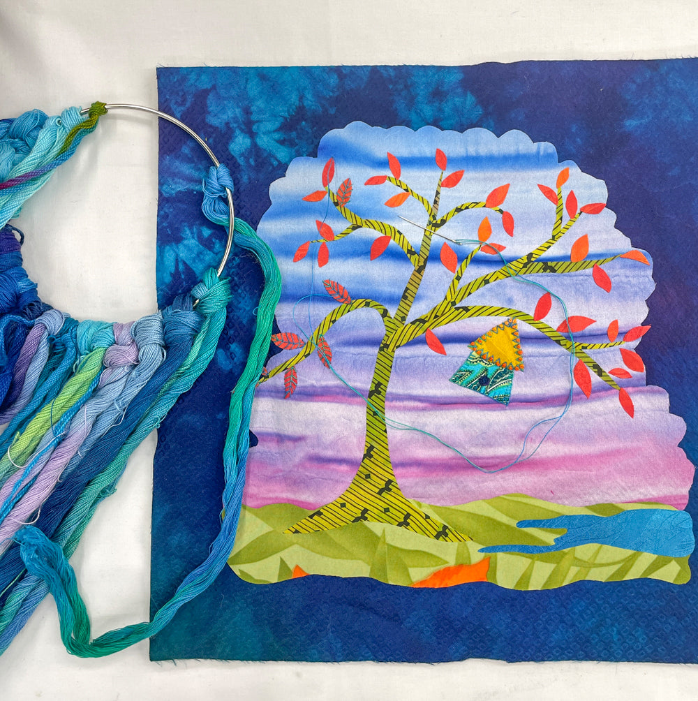 A vibrant quilt square from Creative Spark’s Silk Art Quilts Made Simple with Stitch Library showcases a tree with red leaves on a landscape of blue, purple, and green, beautifully illustrating silk art quilt techniques. Embroidery floss and a hoop rest on the left side, partially attached to the piece, emphasizing the detailed hand embroidery featured in this inspiring project.