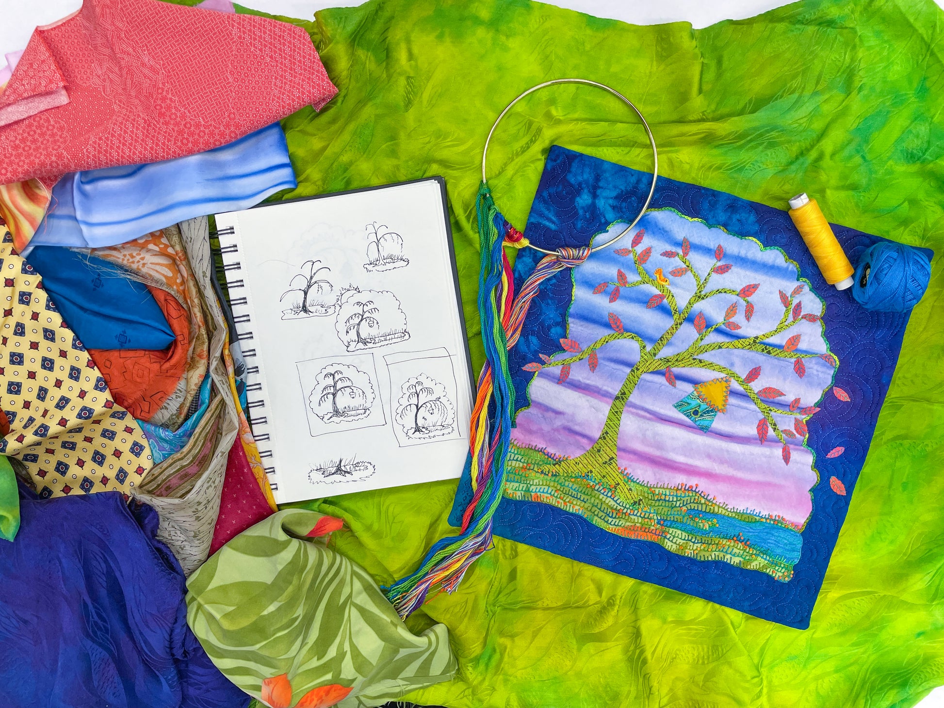 Silk Art Quilts Made Simple with Stitch Library by Creative Spark is displayed alongside colorful fabrics, a sketchbook featuring tree drawings, an embroidery hoop with a hand-embroidered tree design, thread spools, and embroidery floss on a green background.