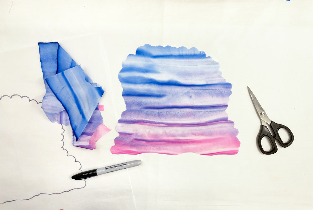 With “Silk Art Quilts Made Simple with Stitch Library” by Creative Spark, you’ll find blue and pink watercolor-dyed fabric pieces, scissors, a black Sharpie, and additional hand-dyed fabrics on a white surface—the perfect elements for crafting your own unique silk art quilt or enhancing your project with detailed hand embroidery.