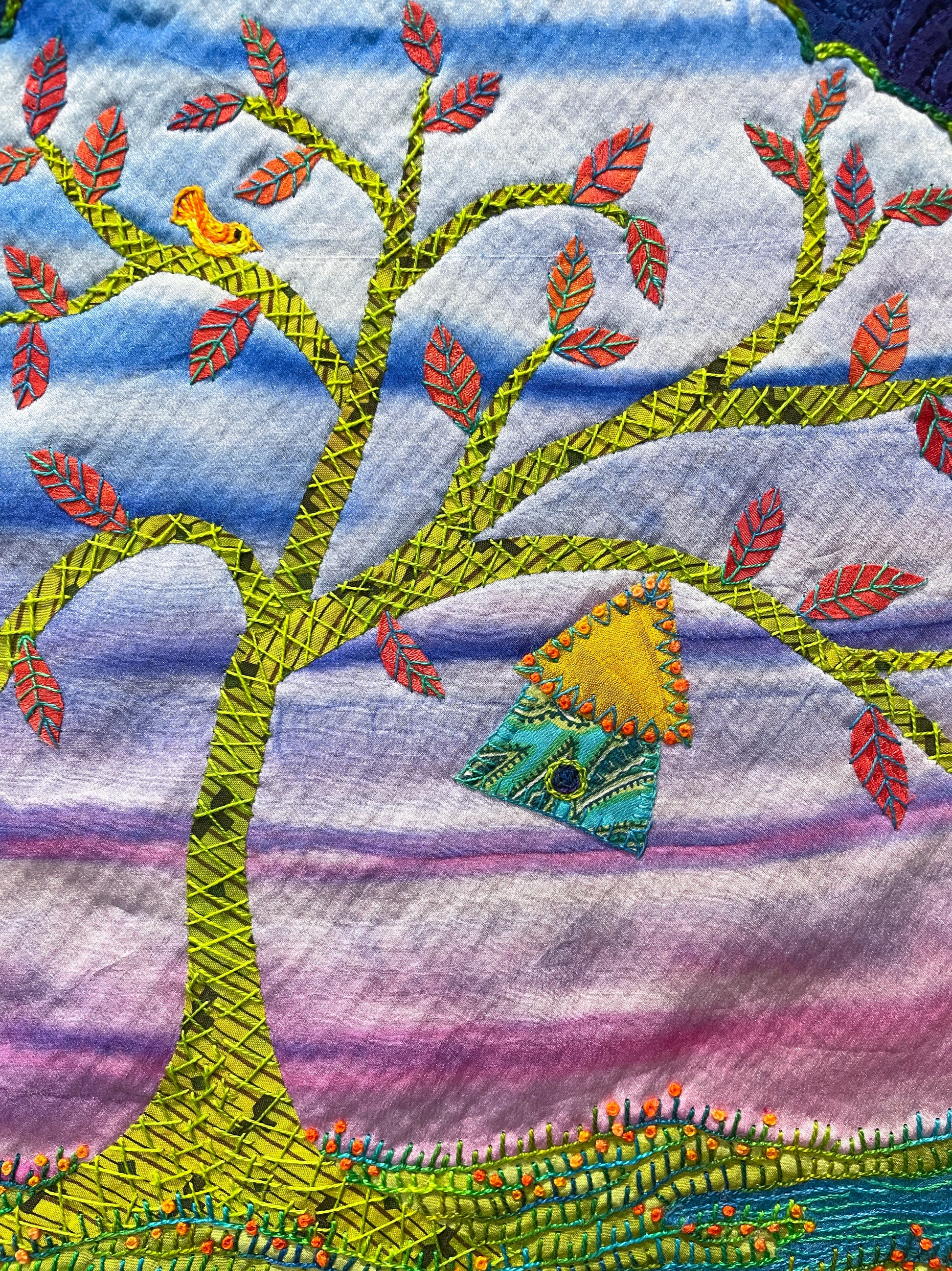 The Creative Spark “Silk Art Quilts Made Simple with Stitch Library” features a vibrant silk art quilt depicting a tree adorned with red-accented leaves, a small bird perched on a branch, and a charming blue birdhouse hanging from another limb, all skillfully brought to life through fusing and delicate hand embroidery set against a vivid purple and blue sky.