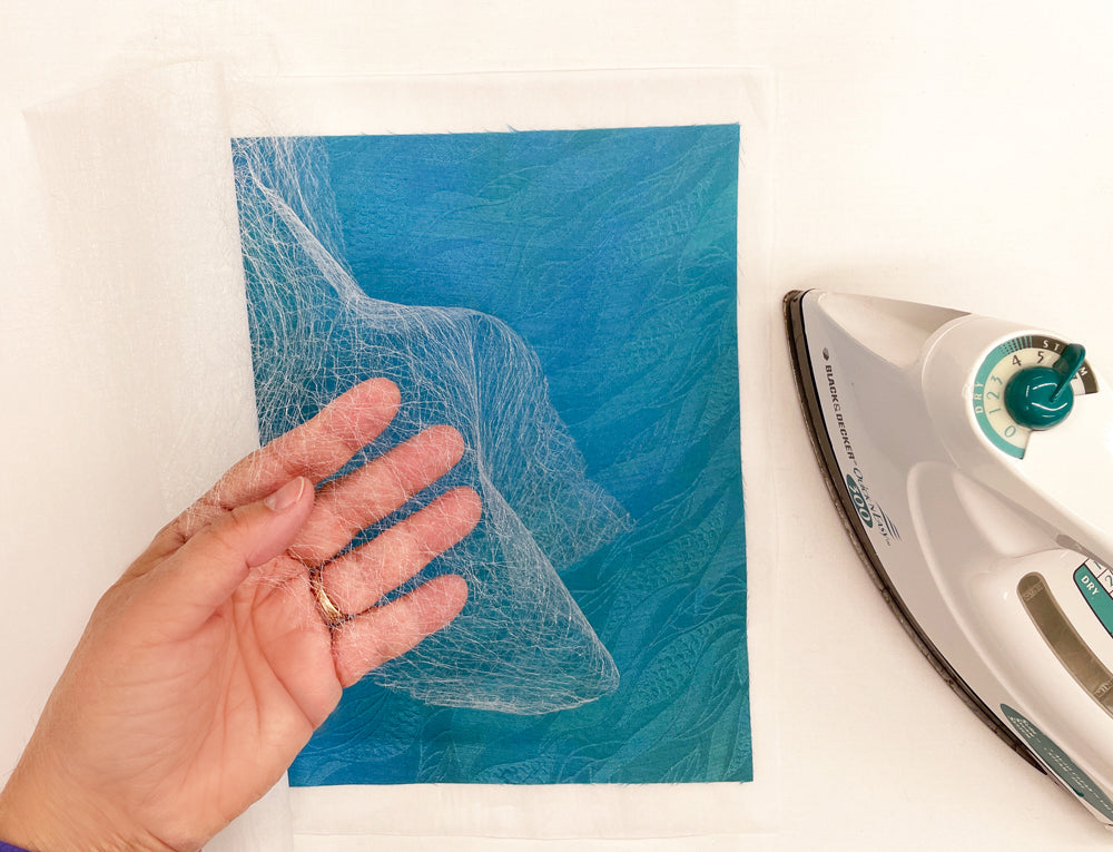 A hand holds thin white mesh over blue fabric art beside a white iron on a table, preparing to fuse materials in the creation of a silk art quilt using Silk Art Quilts Made Simple with Stitch Library by Creative Spark.