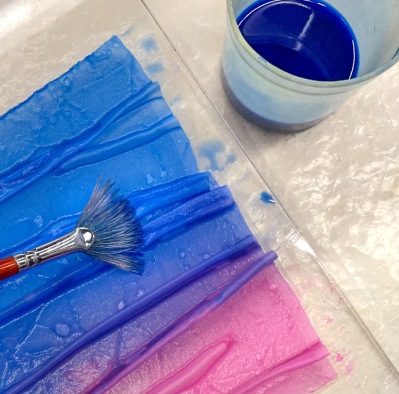 A fan brush rests on textured blue and pink paint strokes beside a cup of blue paint on a light, slightly reflective surface, capturing the layered creativity and delicate detail featured in Creative Spark’s *Silk Art Quilts Made Simple with Stitch Library*.