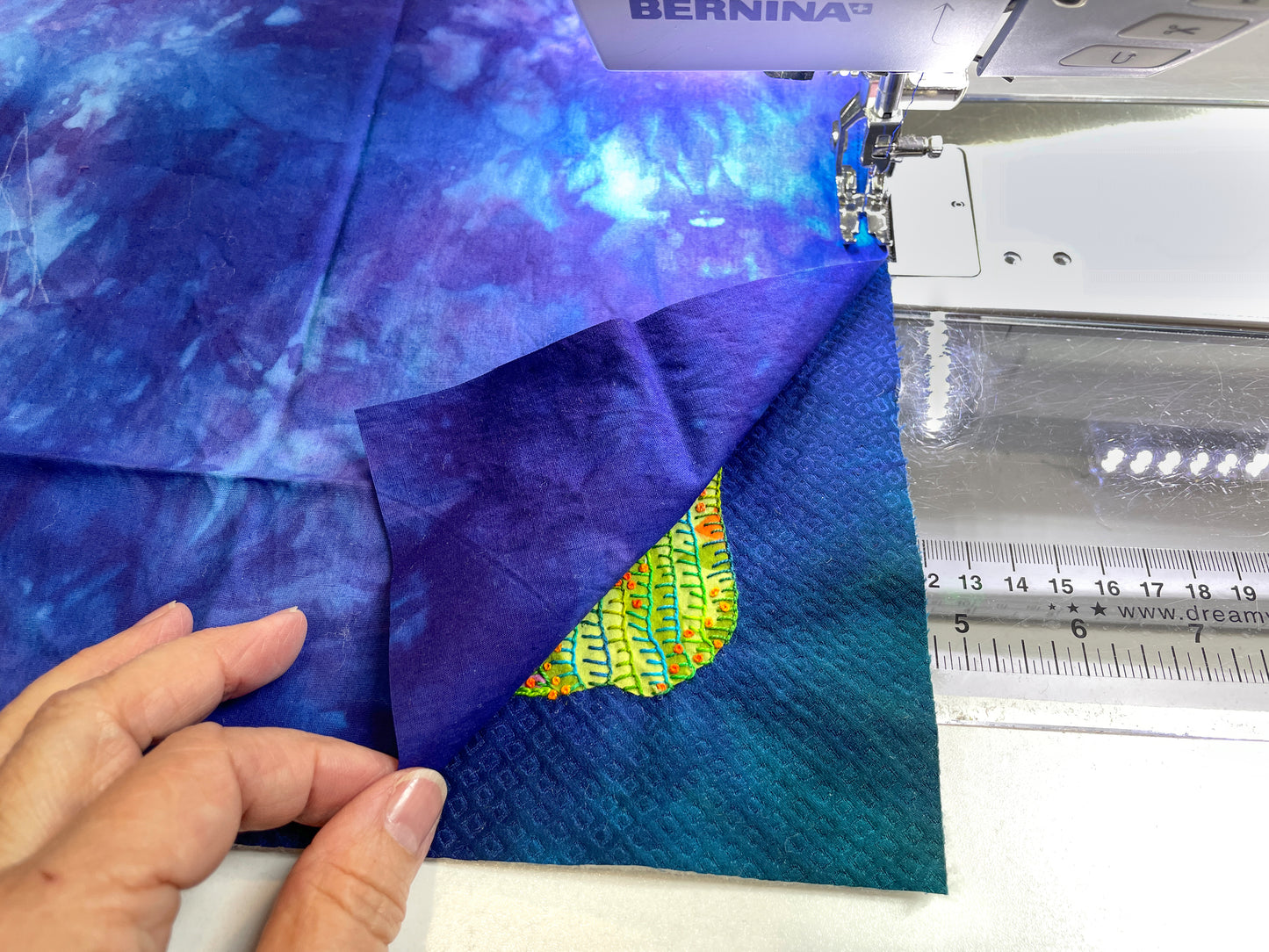 A hand lifts the corner of blue fabric to reveal a colorful, hand-embroidered leaf patch beneath, next to a sewing machine with a ruler and needle visible. The Silk Art Quilts Made Simple with Stitch Library kit by Creative Spark features expert fusing techniques for added detail and texture.