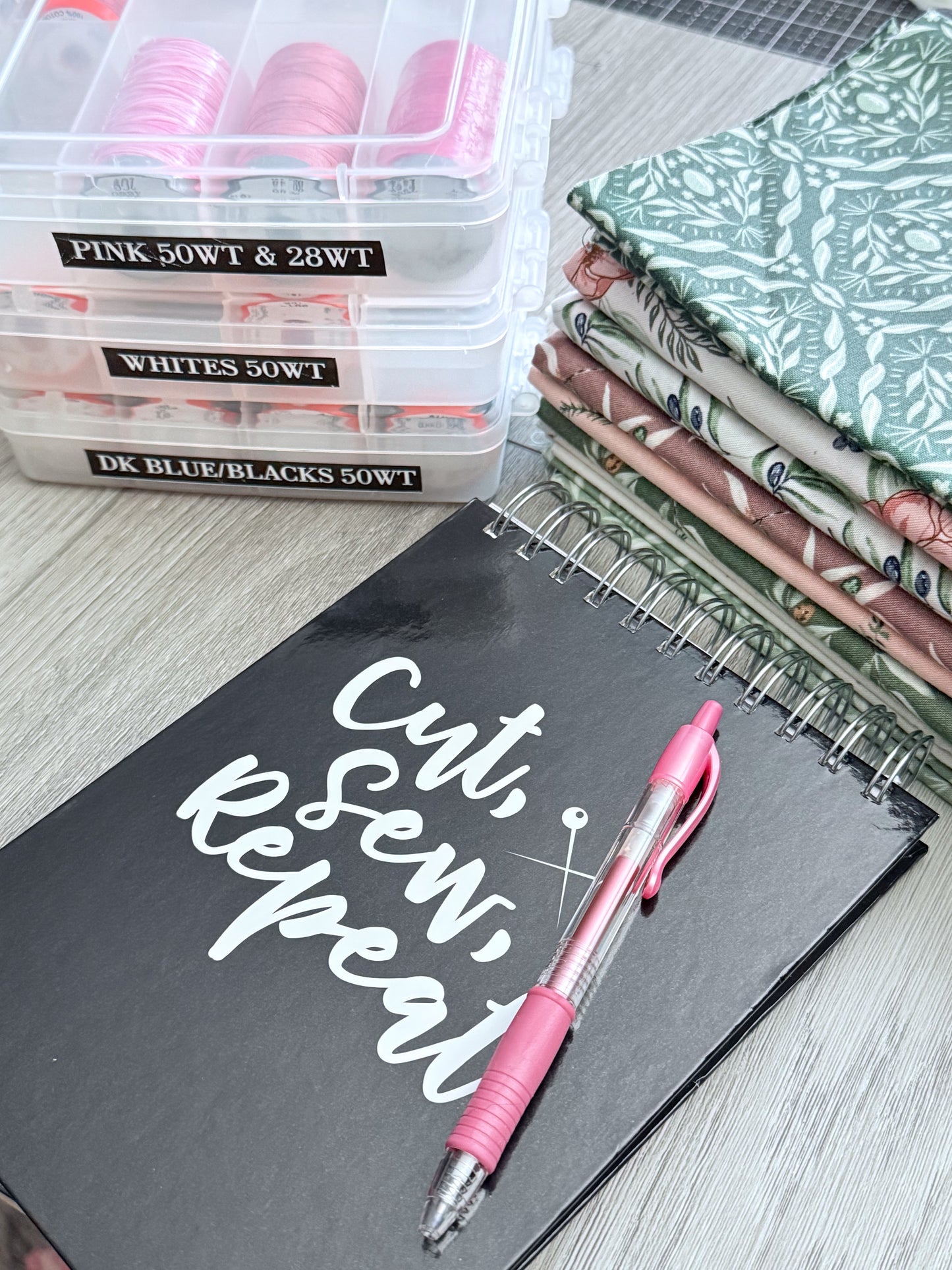 Arranged on a wooden surface are essentials for creative organization: the Creative Spark “Organize Your Creative Space: A Step-by-Step Craft Room Reset” notebook, a pink pen, stacks of patterned fabric, and color-coded plastic thread cases—perfect inspiration for thoughtful sewing supplies storage and craft room organization.