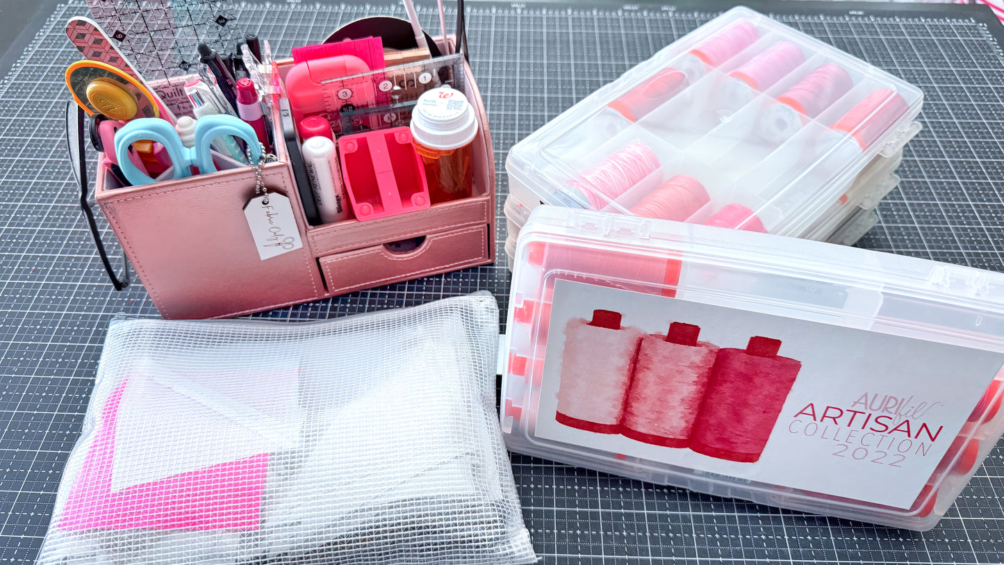 The Creative Spark "Organize Your Creative Space: A Step-by-Step Craft Room Reset" features a pink organizer with sewing tools, clear boxes filled with pink and white thread, and a mesh pouch containing fabric pieces—all neatly arranged on a black cutting mat—offering the perfect solution for quilting organization and keeping your sewing supplies storage neat and accessible.