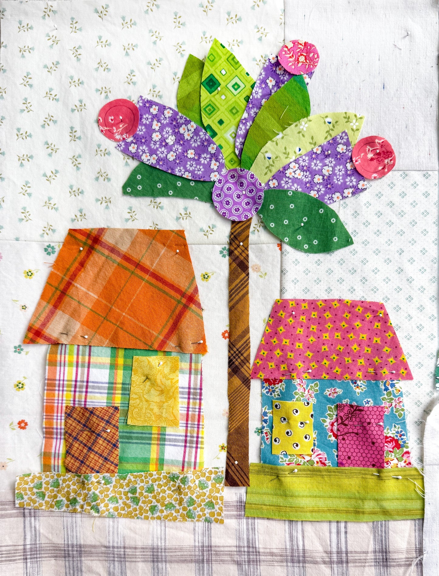 Buttonwood Village: Build an Adventure Block of the Month by Creative Spark features whimsical houses, a tall tree with green and purple leaves, and pink flowers on a light patterned background—perfect for your next appliqué project!.