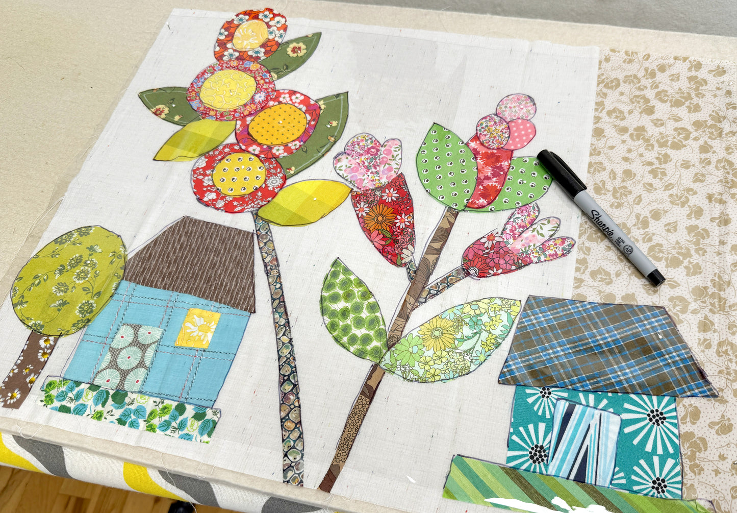 A fabric collage for Creative Spark’s Buttonwood Village: Build an Adventure Block of the Month shows colorful flowers and whimsical houses on white cloth, with a black marker on a beige patterned surface.