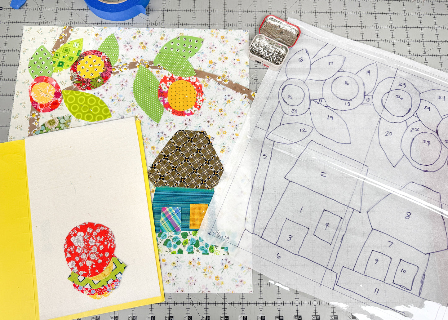 A crafting workspace features Creative Spark's Buttonwood Village: Build an Adventure Block of the Month, with whimsical house appliqué art, a yellow notebook with red floral appliqué, and a clear folder with the hand-drawn pattern.