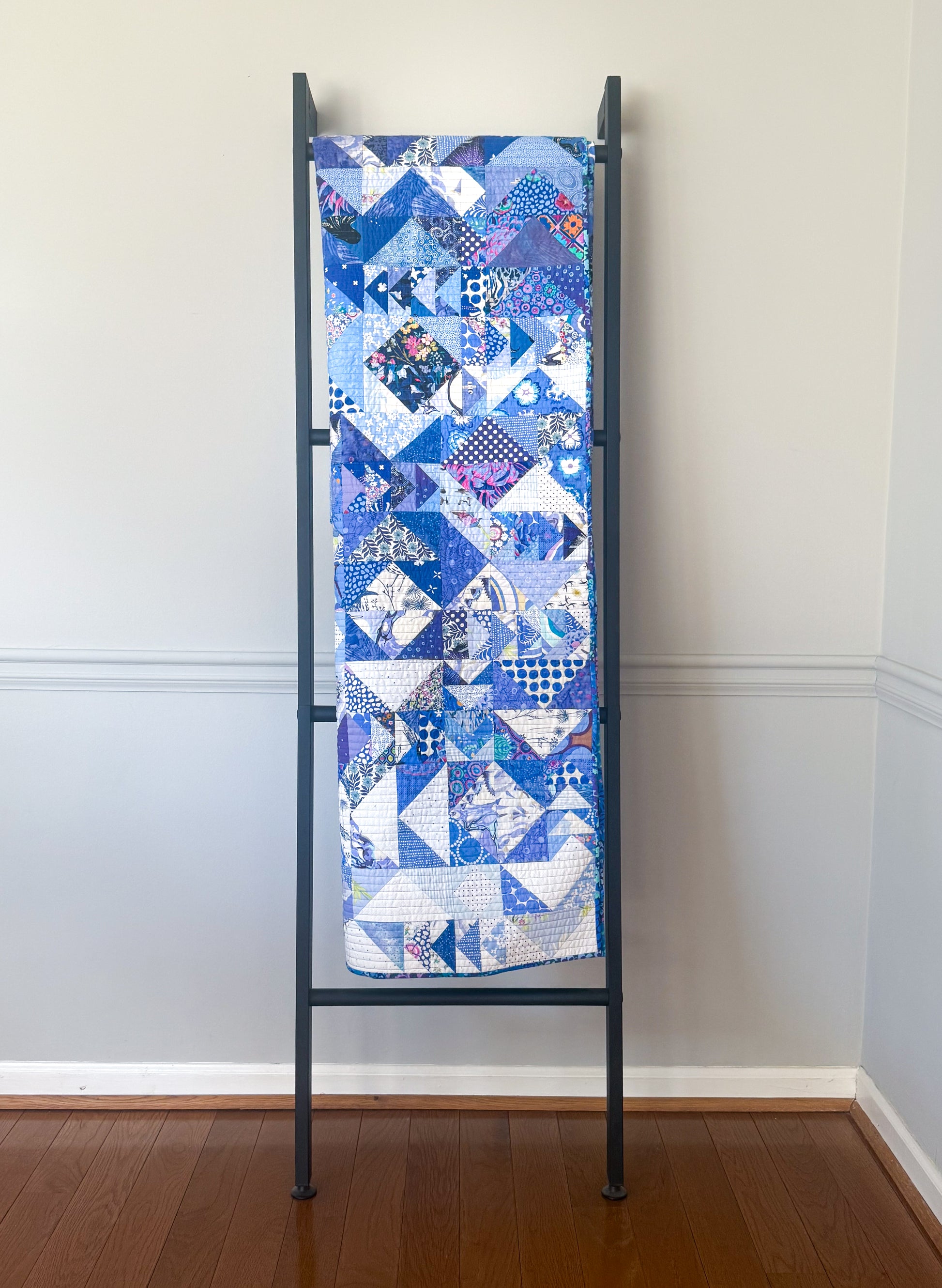 The Misunderstood Quilt: Blend Color, Value & Precision by Creative Spark, featuring blue and white geometric patterns, is draped over a black ladder rack against a light gray wall and white wood floor.