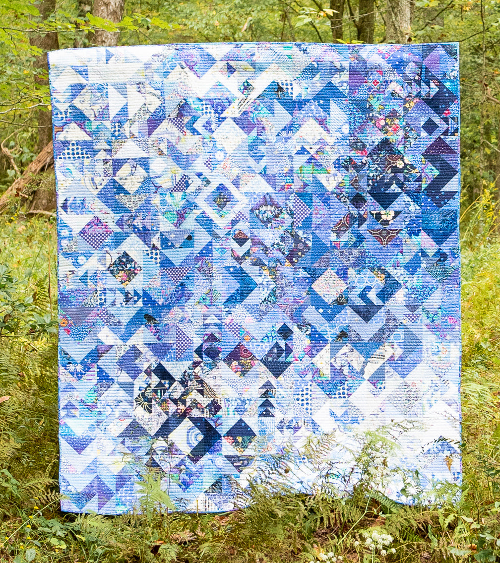 The Misunderstood Quilt: Blend Color, Value & Precision by Creative Spark—a large blue and white geometric patchwork—hangs vertically outdoors among green foliage and trees, showcasing expert precision piecing.