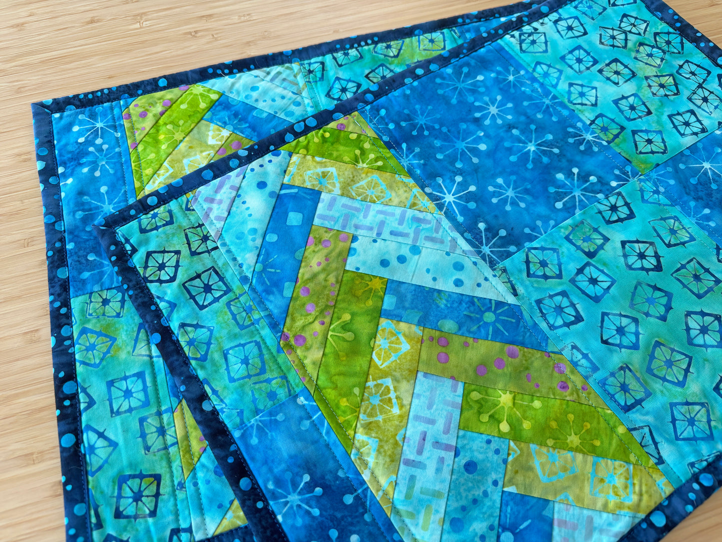 Two "Patchwork Braids 101 Basics" quilted placemats by Creative Spark, in blue and green geometric and snowflake patterns, overlap on a light wood surface.