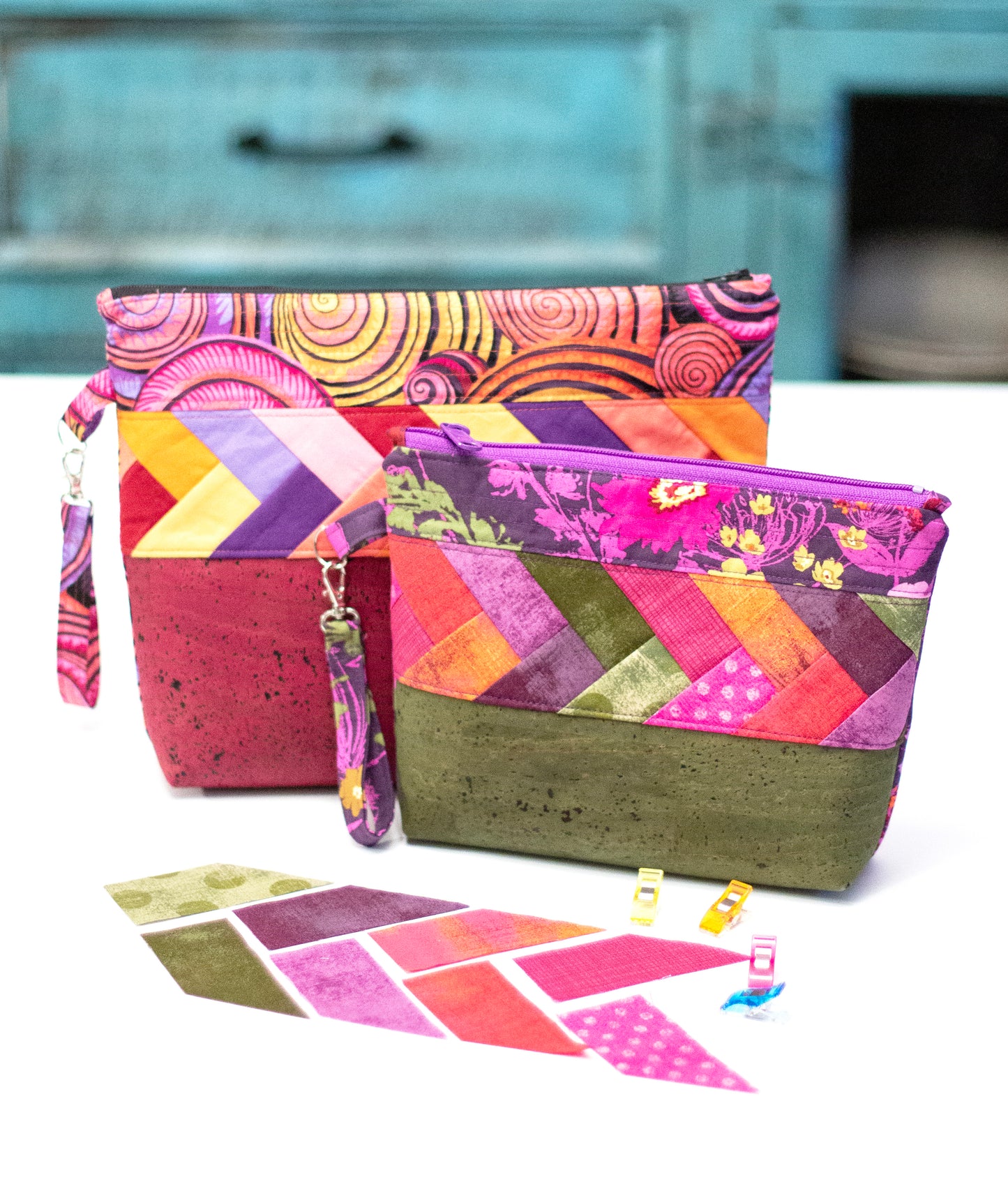 Two colorful zippered pouches made with Patchwork Braids 101 Basics by Creative Spark feature geometric patchwork and playful braids in pink, purple, orange, and green. They’re shown with matching fabric pieces and sewing clips on a white surface. Blue furniture is blurred behind.