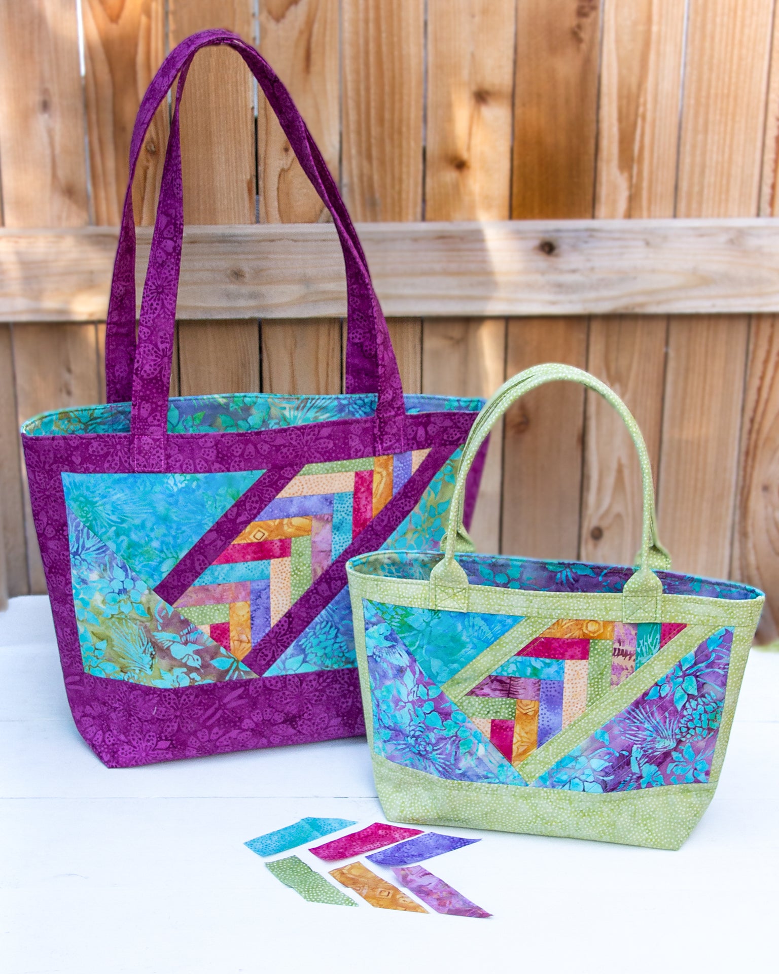 Two quilted tote bags made with Patchwork Braids 101 Basics by Creative Spark, featuring chevron designs and bold braids in purple, green, and blue, are displayed on a white surface before a wooden fence with matching fabric strips in front.