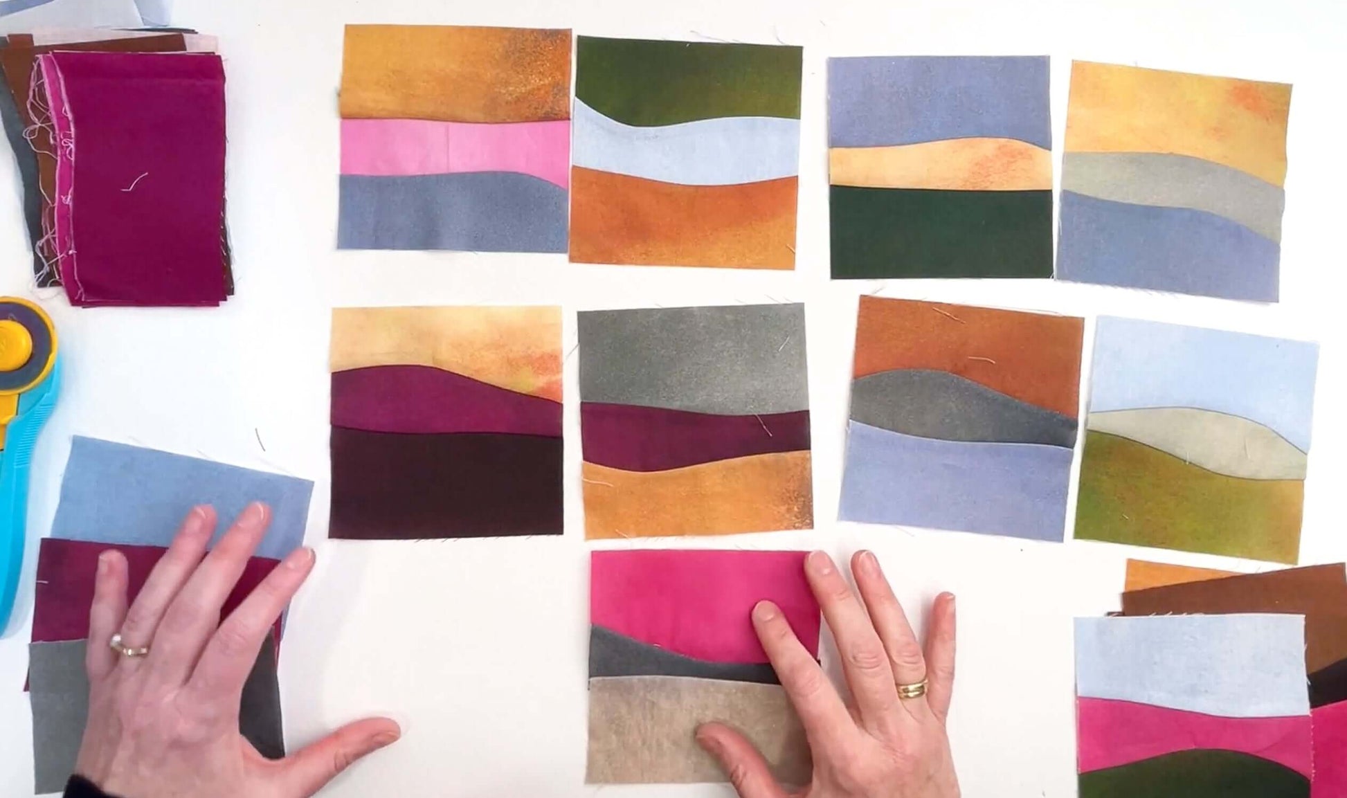 A person arranges vibrant fabric squares with abstract, wavy designs—ideal for scenic quilts or improv landscapes using Creative Spark's Landscapes as Building Blocks: Quilt the Places You Love with Easy Improv. Stacks of fabric and a rotary cutter sit to the left.