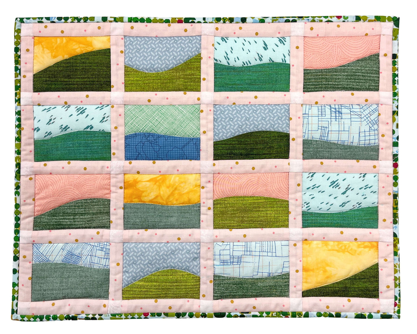 The Creative Spark "Landscapes as Building Blocks: Quilt the Places You Love with Easy Improv" kit features a colorful quilt of sixteen abstract landscape blocks—hills and skies in pink, yellow, and blue—framed by light pink sashing with gold dots.