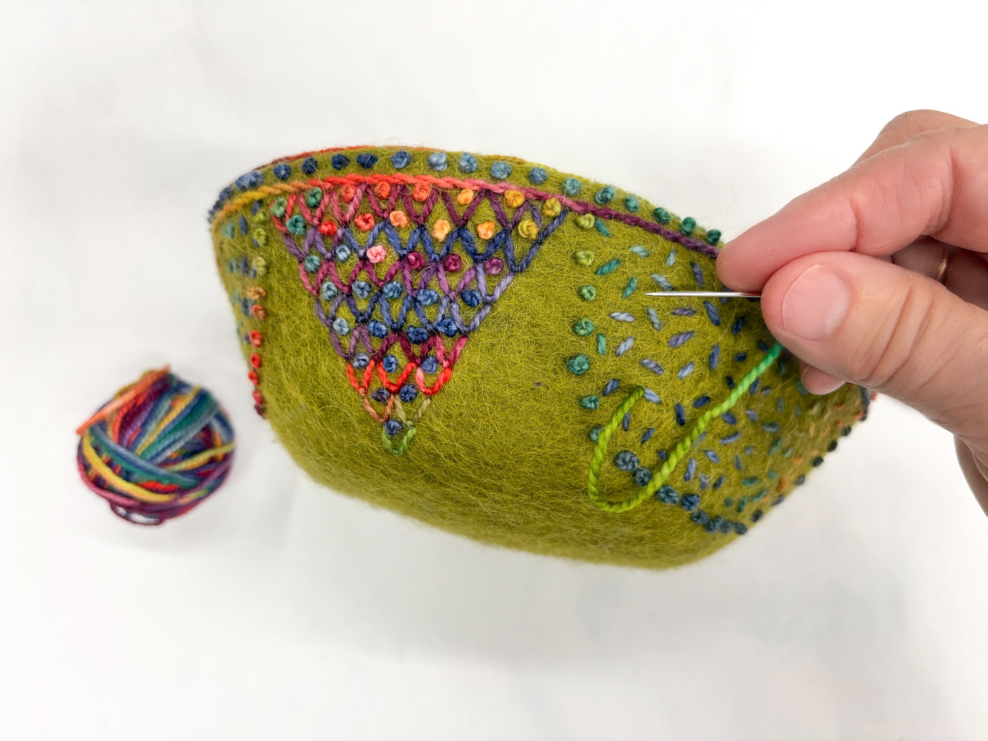 A hand stitches vibrant embroidery onto a green felt bowl in "Embroidery in the Round: Learn Stitching & Embellishment on Felt Bowls" by Creative Spark, with a colorful yarn ball nearby on a white surface.