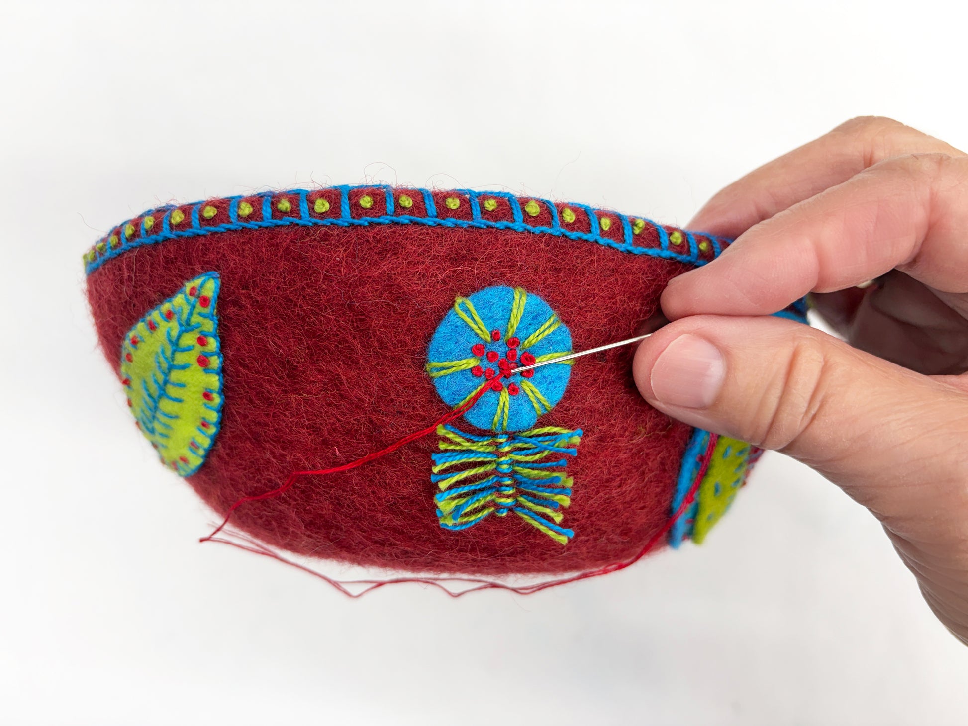 The Embroidery in the Round kit by Creative Spark teaches you to stitch colorful blue, green, and yellow designs onto red felt bowls using vibrant appliqué and embellishment techniques.