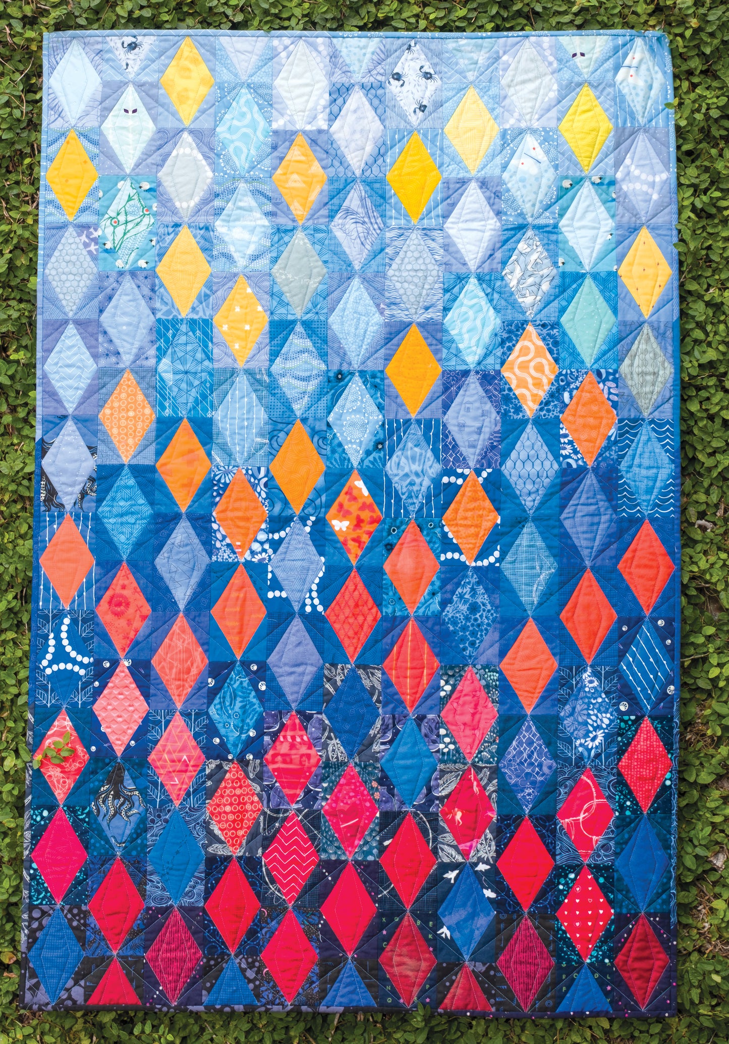 Experience the vibrant design of the *Using Scraps in Modern Quilts: Design Lab* by Creative Spark—a lively quilt showcasing diamond patterns in blue, yellow, orange, and red, artfully displayed on lush green grass. The inventive use of negative space enhances its modern aesthetic and brings each color to life.